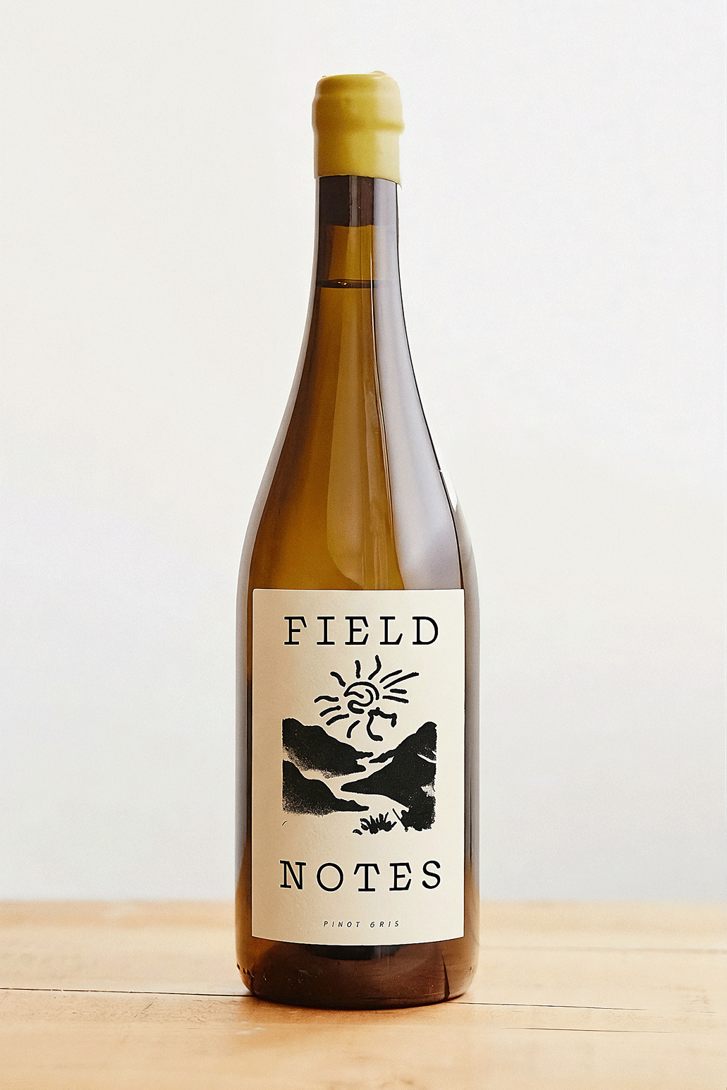 A bottle of Field Notes Pinot Gris wine with a label featuring a mountain landscape and a sun, placed on a wooden surface against a plain background.