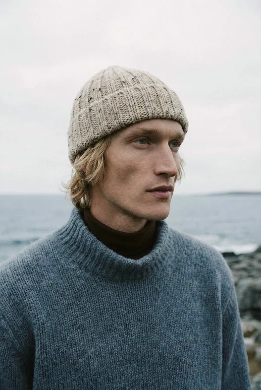 A young man with blond hair wearing a beige knit beanie and a gray sweater standing outdoors by the water on a cloudy day.