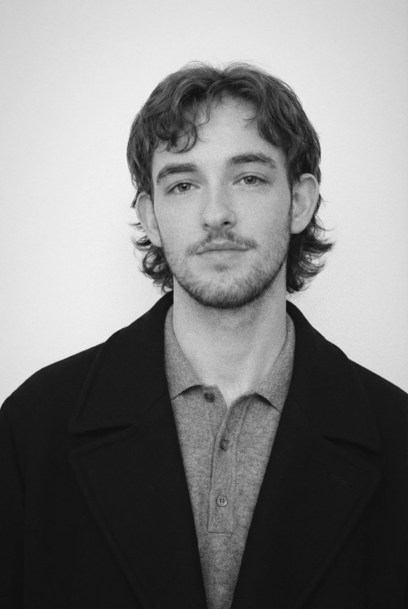 Black and white portrait of a young man with wavy hair, facial hair, and a serious expression, wearing a collared shirt and a coat, against a plain background.