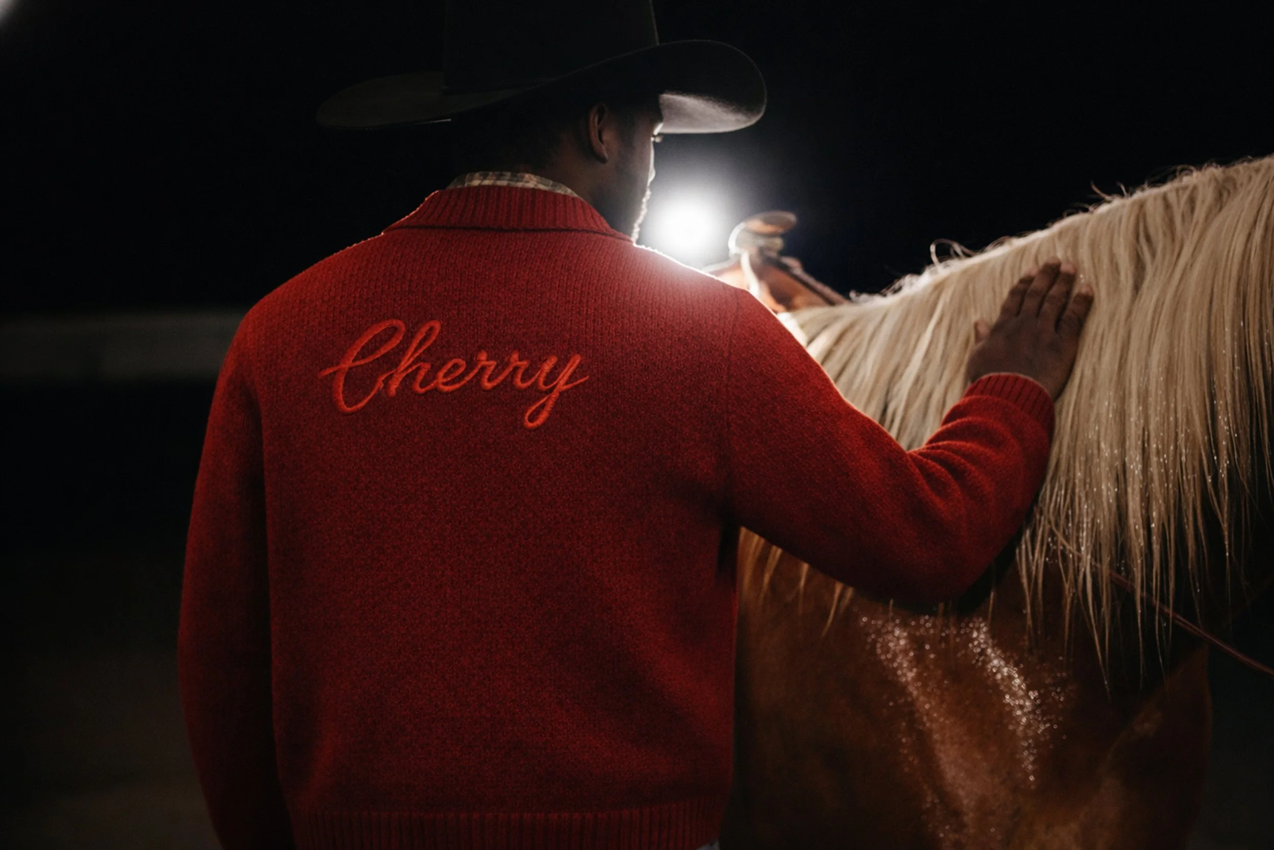 A man wearing a red sweater with the name "Cherry" embroidered on the back, a black hat, and a plaid shirt underneath, touching the mane of a white horse in a dark setting with backlighting.