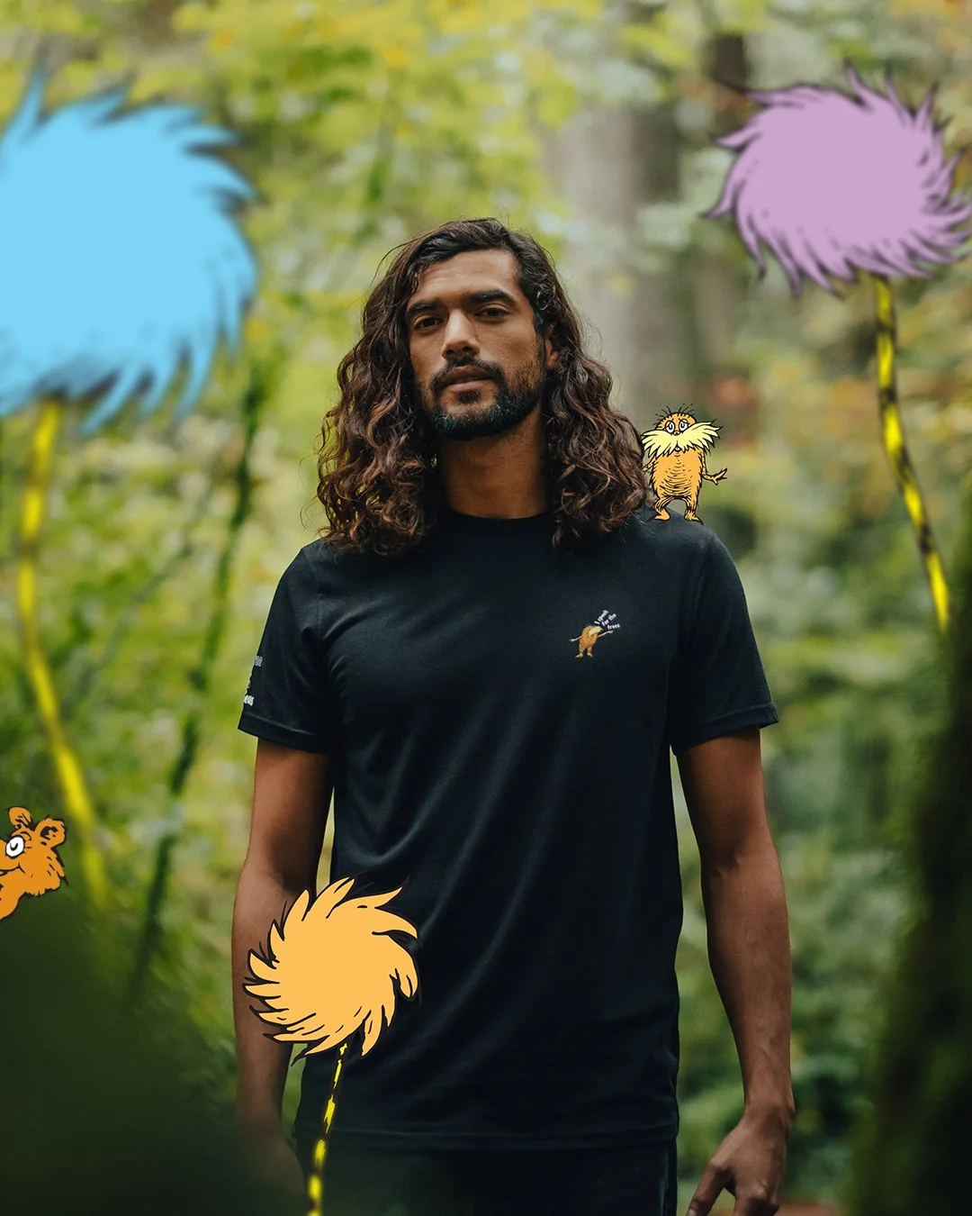 A man with long, curly dark hair and a beard standing in a forest surrounded by cartoonish, colorful, fluffy animals and plants.