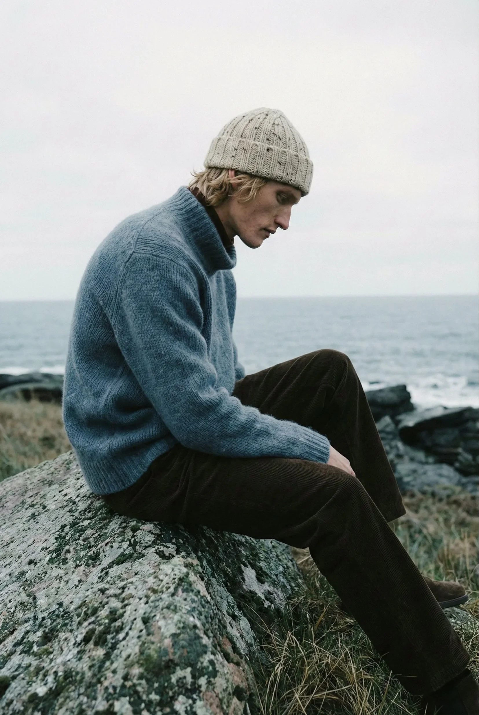 A young man with blond wavy hair, wearing a beige knit beanie and a blue sweater, sitting on a rock near the ocean, looking down in a contemplative pose.