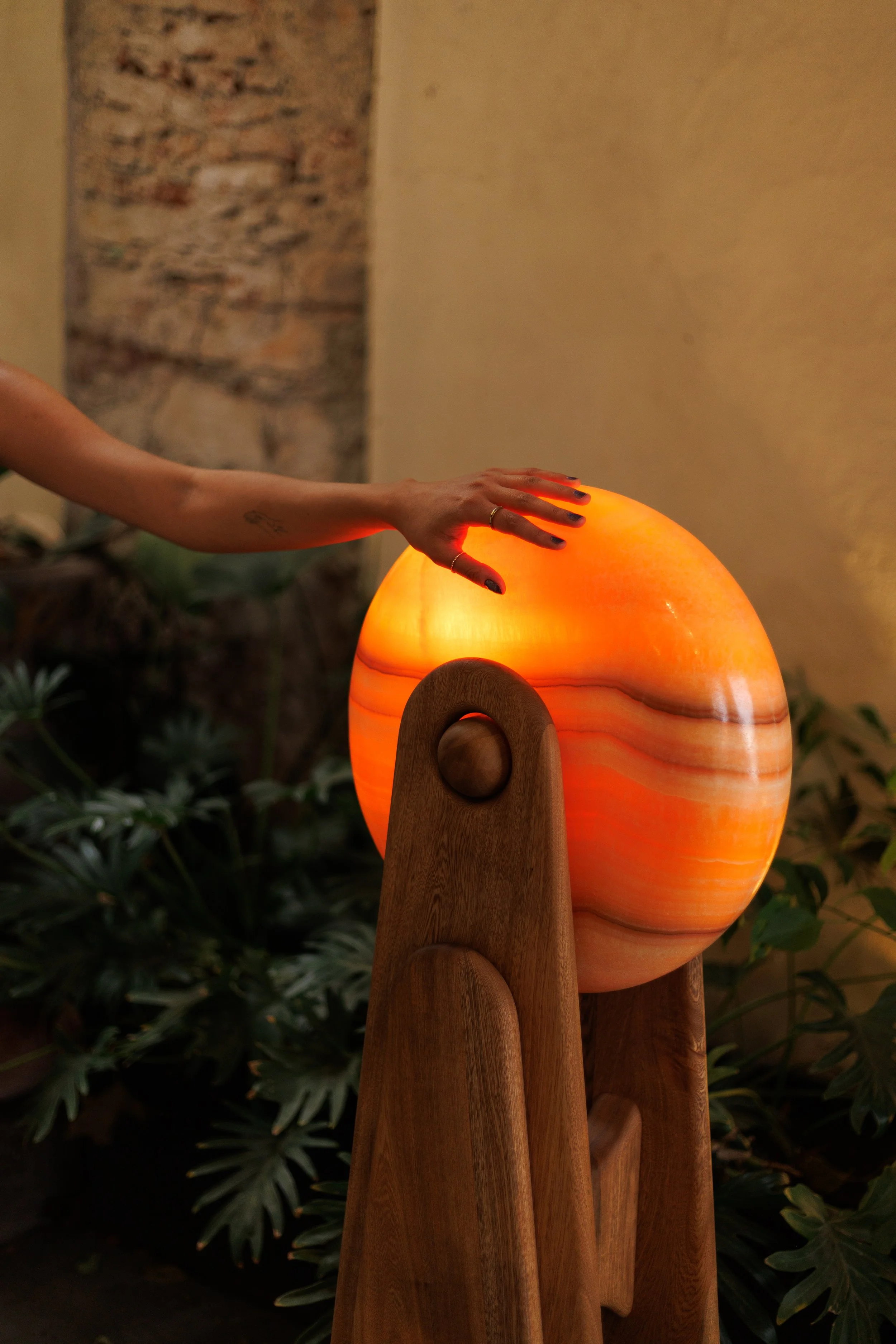 A person's hand with black painted nails is reaching out to touch a glowing orange spherical lamp, which is mounted on a wooden stand. The background includes a stone wall and some leafy green plants.