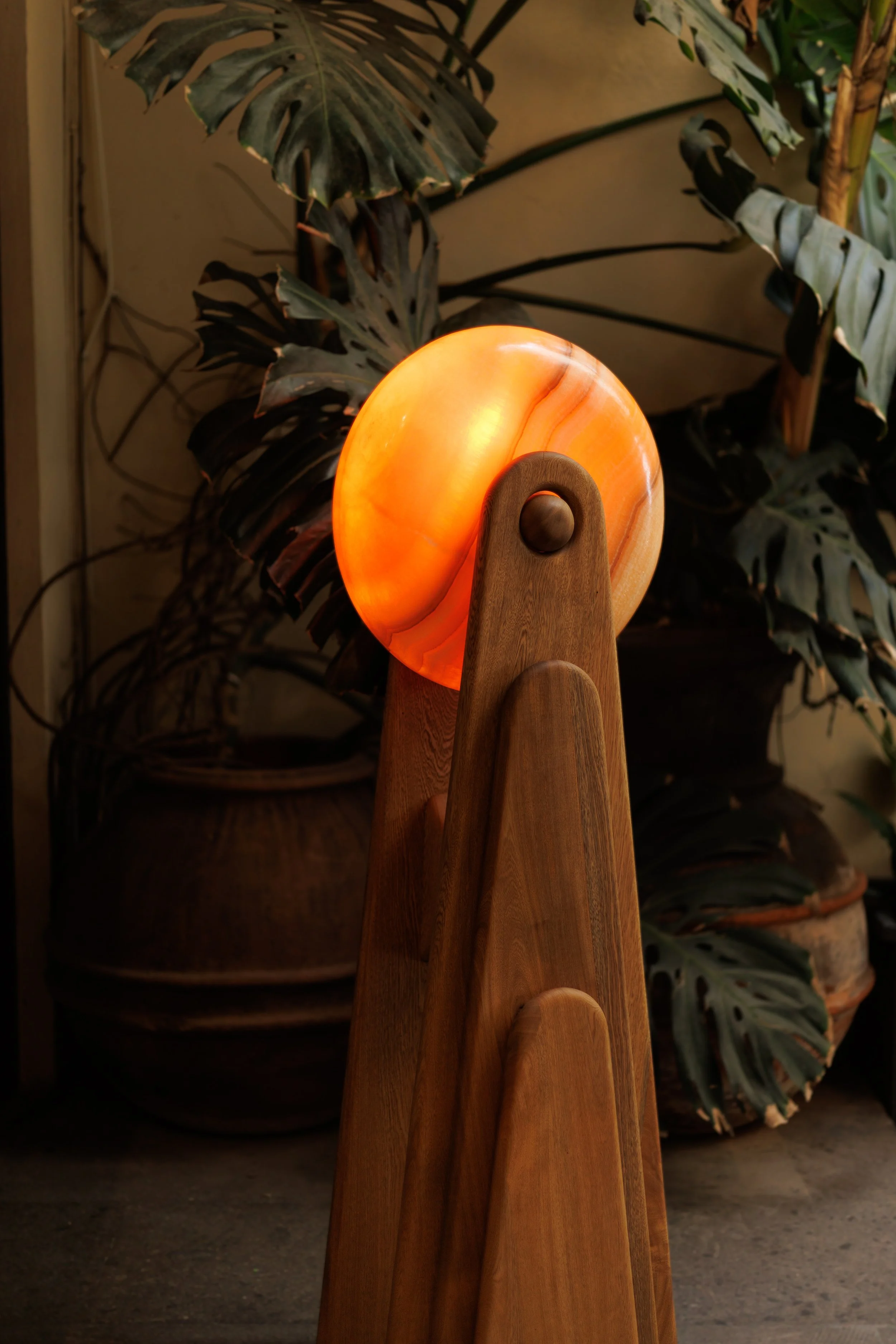 A wooden sculpture with a glowing orange orb on top, set against a background of large green tropical leaves.