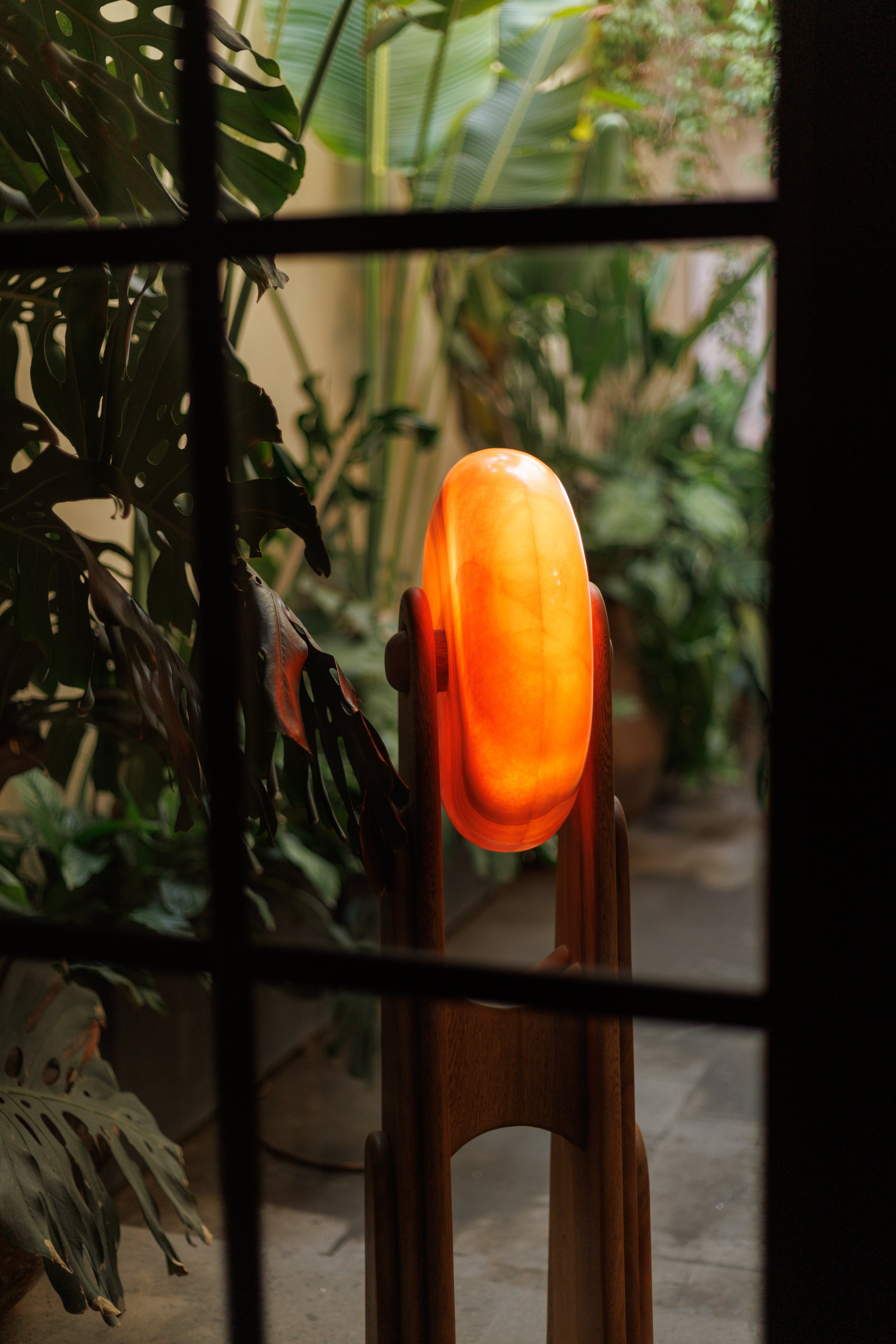 A glowing orange lamp on a wooden stand is seen through a window with black bars, surrounded by green indoor plants.