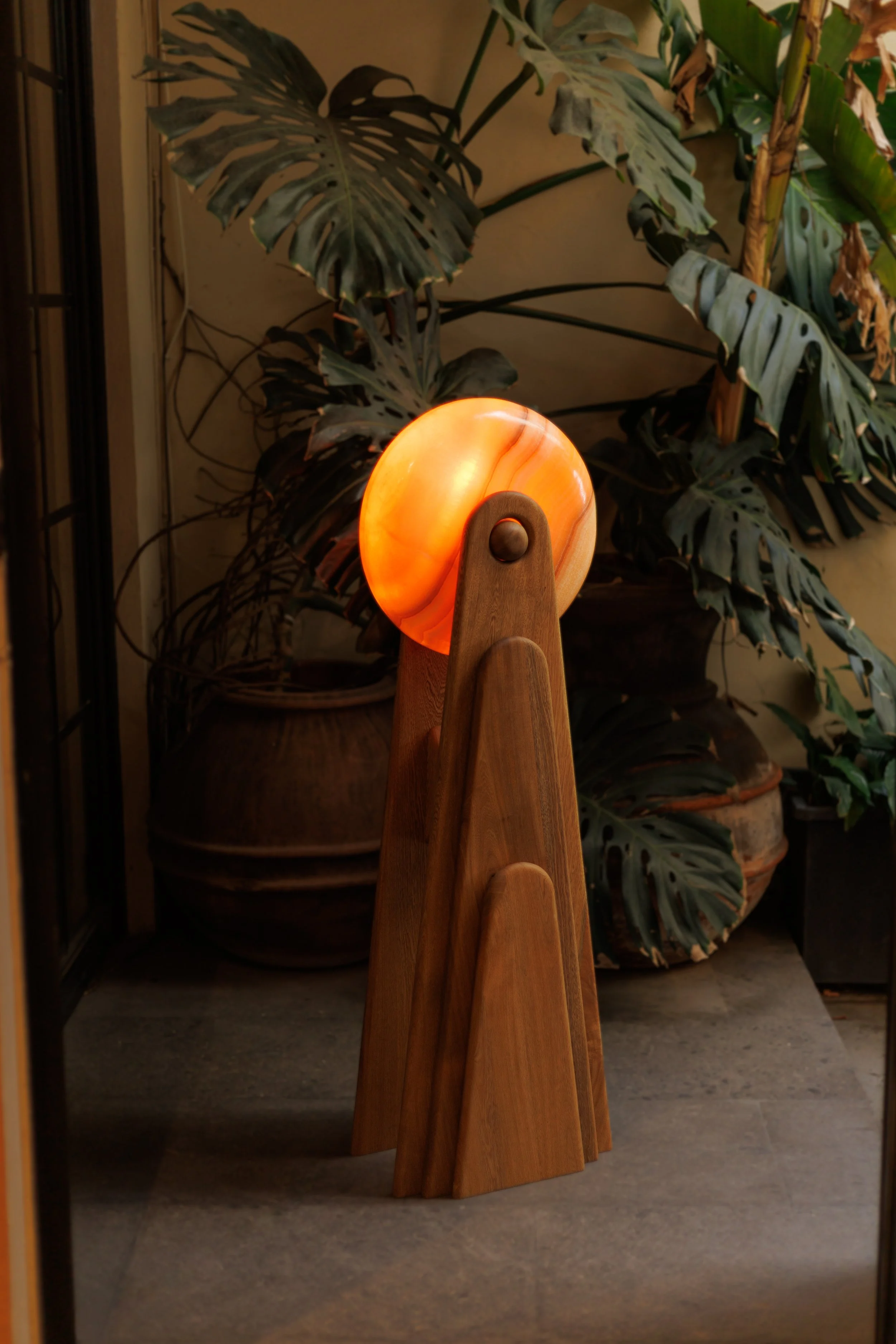 A decorative lamp with an orange onyx sphere on top, mounted on a wooden stand, placed on a floor surrounded by large potted plants with broad leaves.