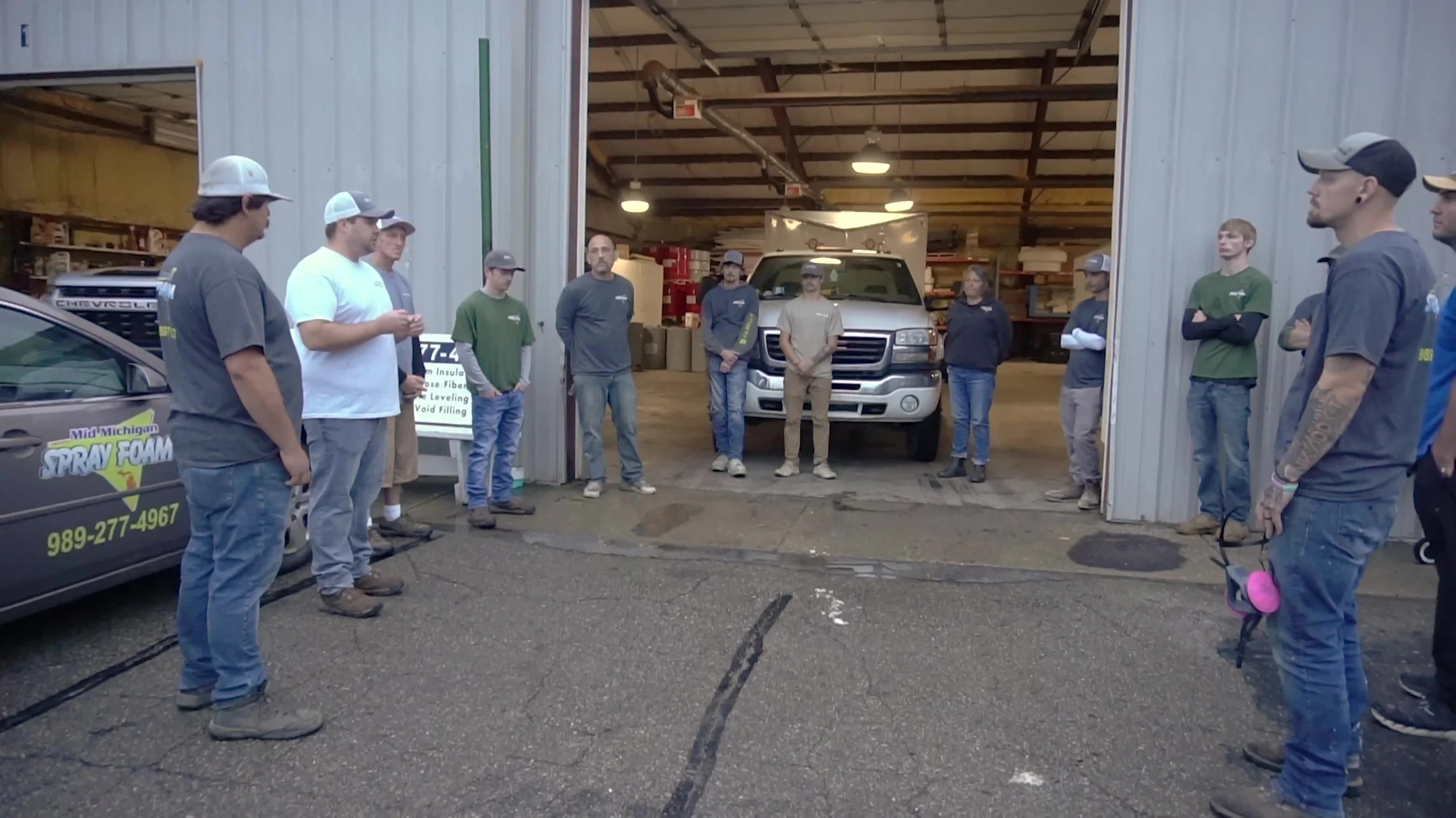 Group of people standing outside a warehouse or workshop, with a truck inside. They appear to be part of a work crew or team meeting.