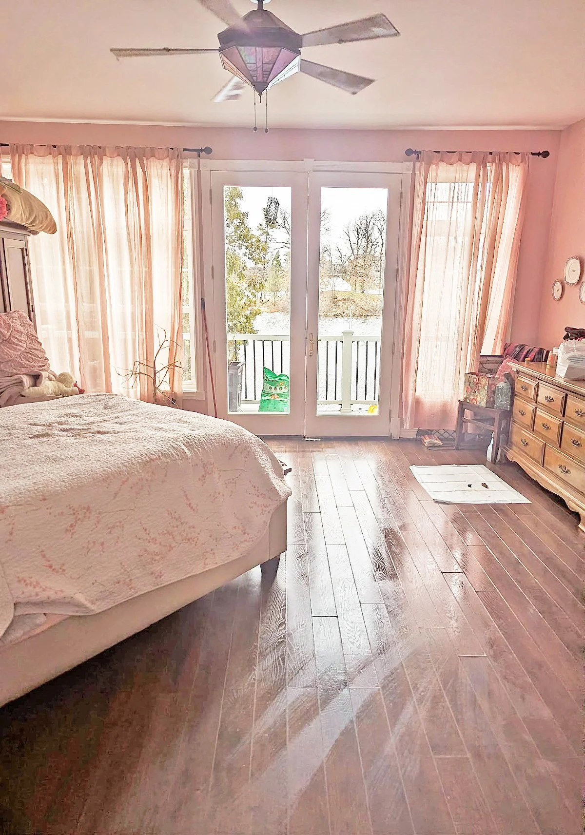 A bedroom with pink walls, a bed with a light-colored quilt, pink curtains, a wooden dresser, and glass double doors leading to a balcony with a view of trees and a river outside.