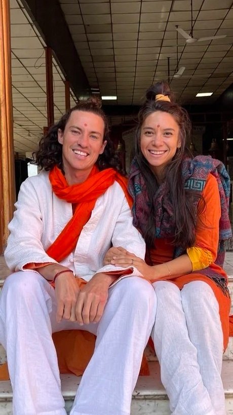 Two women sitting together indoors, smiling, wearing traditional Indian clothing, with one in white and orange and the other in colorful attire.