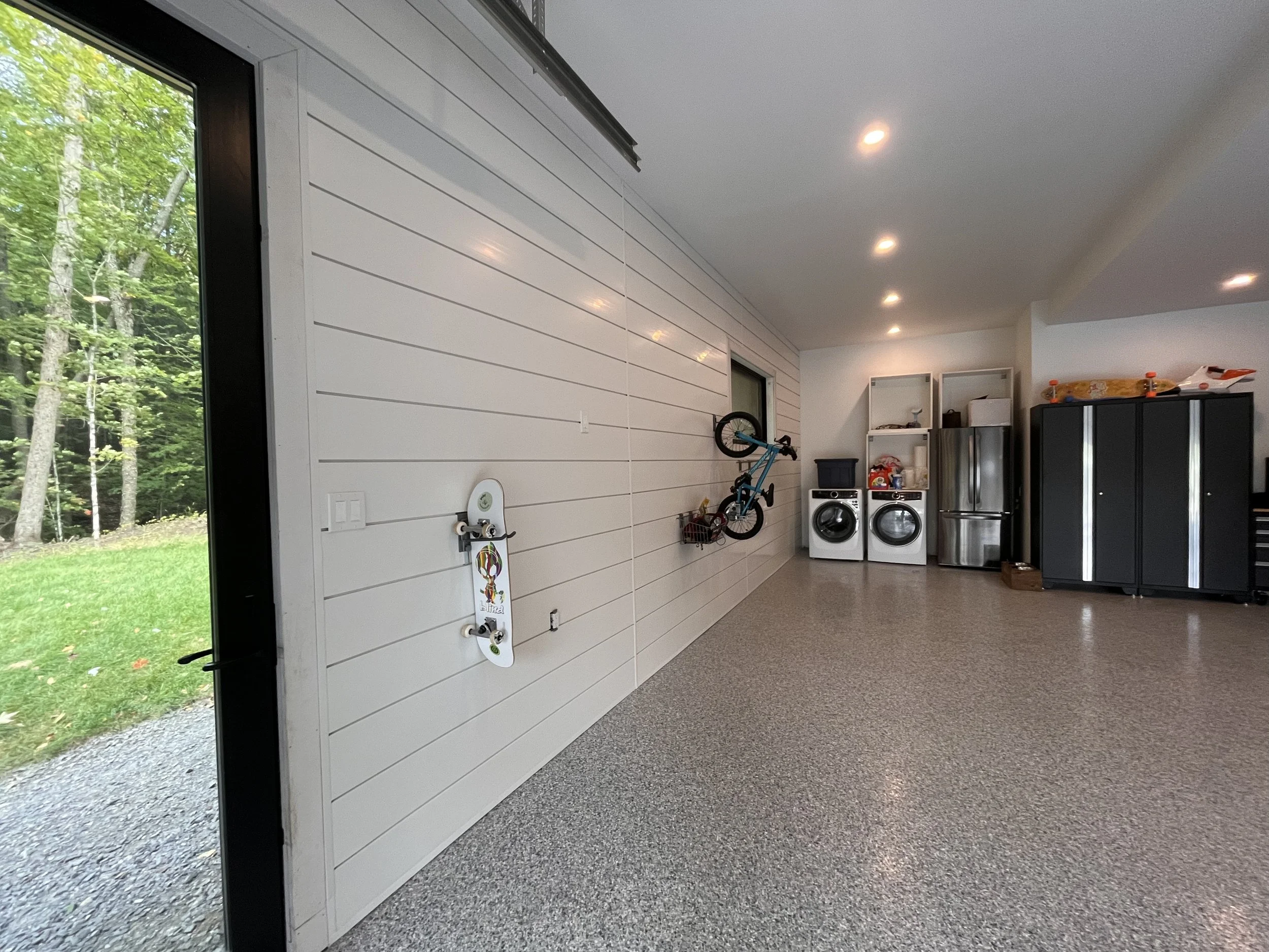 Garage with white panel walls, gray flooring, and ceiling. Inside are a blue bicycle mounted on the wall, laundry washer and dryer, black storage cabinets, and open shelves with various items. A glass door shows greenery outside.