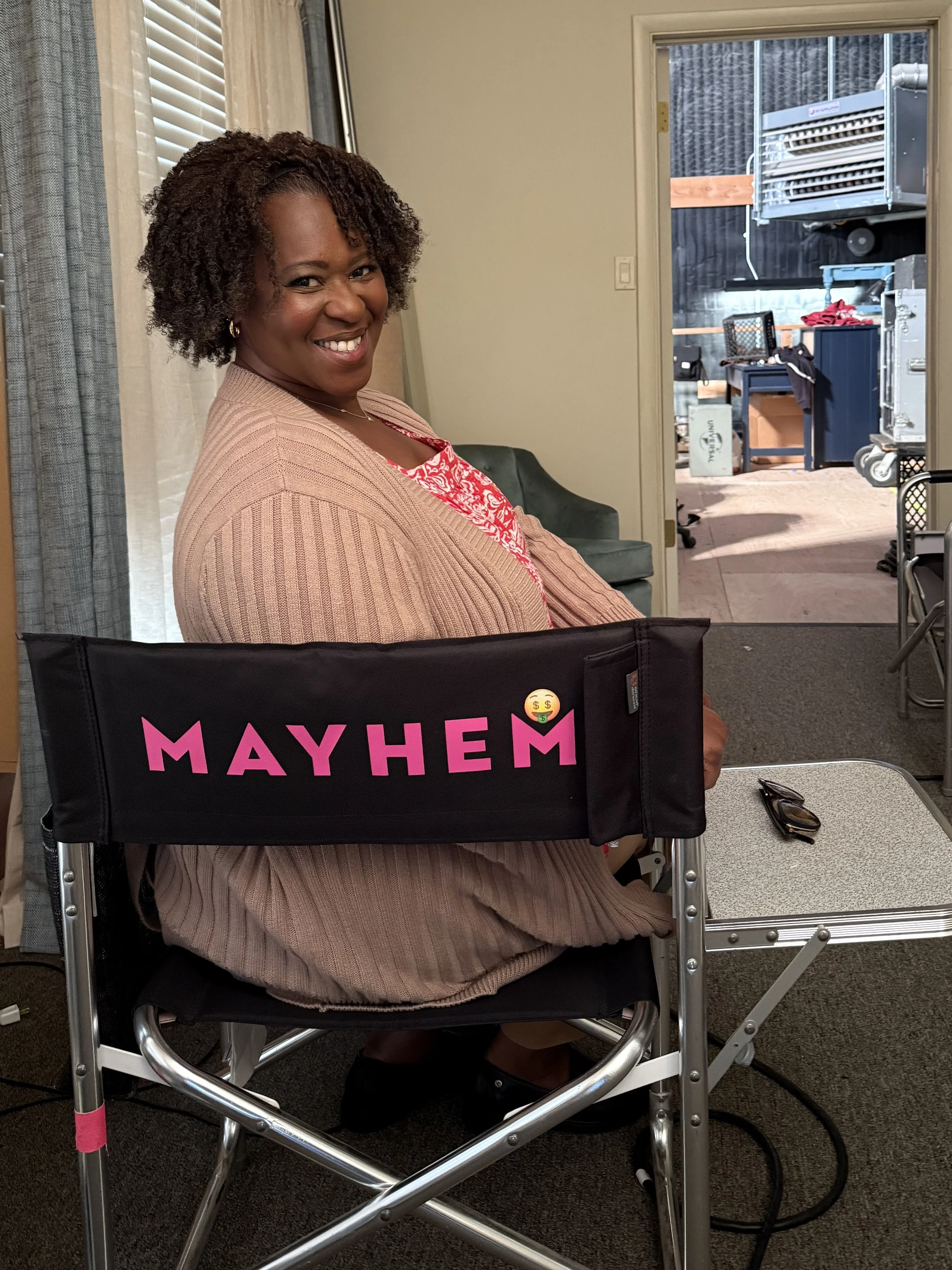 A smiling actor, Treisa Gary is sitting on a black cast chair with the words 'MAYHEM' written on it in pink and a smiley face with dollar signs emoji.