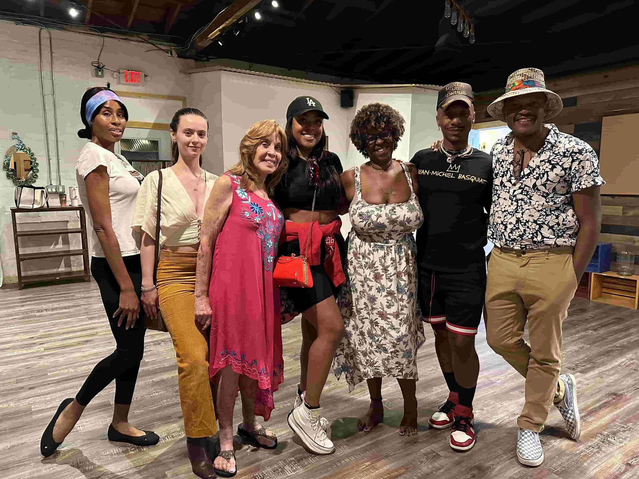Group of seven people smiling indoors, standing on a wooden floor. They are dressed casually, and some wear hats. The setting appears to be a room with curtains and wooden accents.