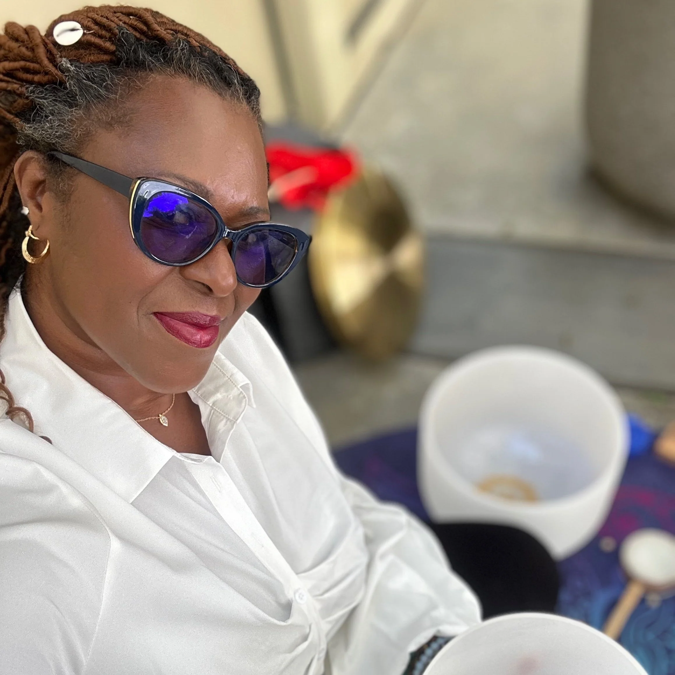 Person wearing sunglasses, seated outdoors with a white shirt, alongside a white bowl and a gong.