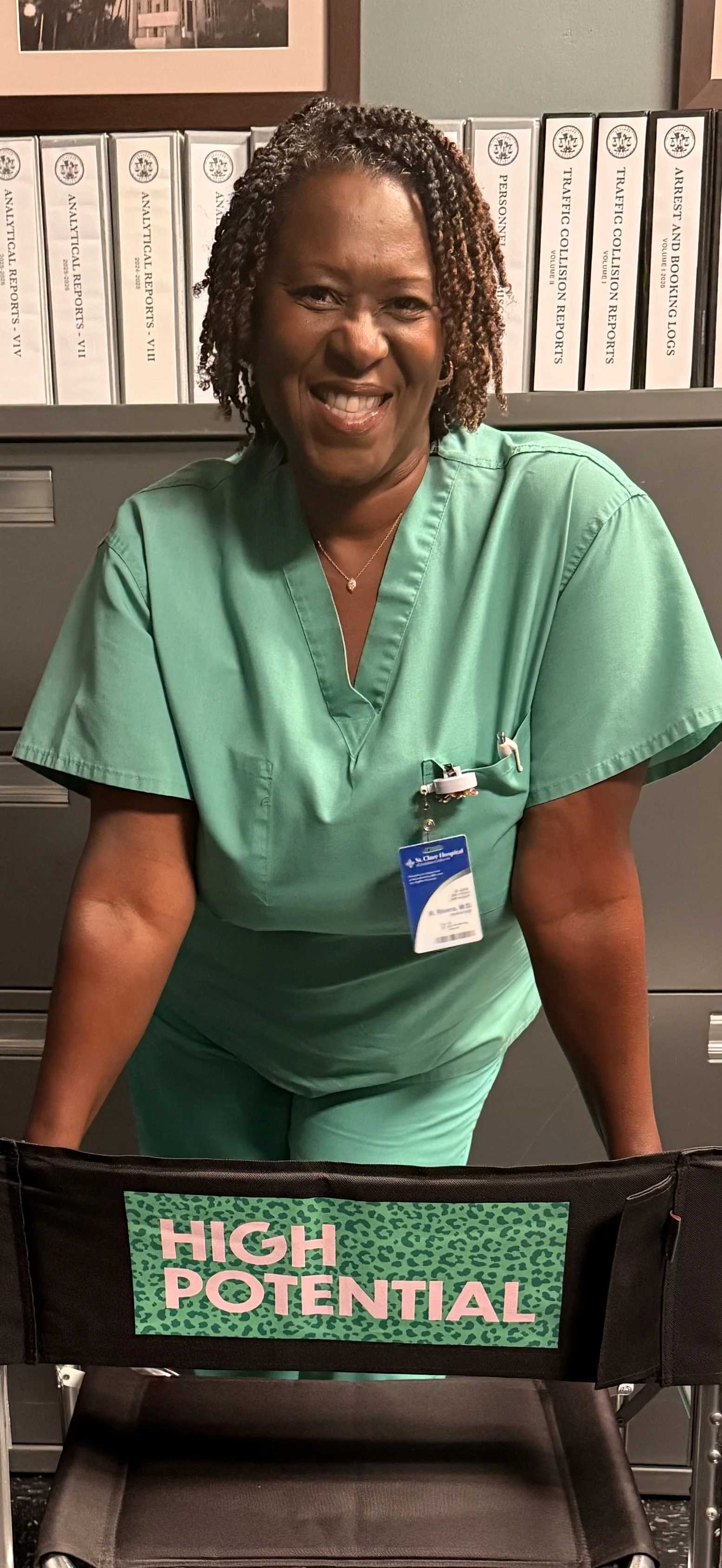 A smiling actor, Treisa Gary, in scrubs stands behind a cast chair on set, that reads 'High Potential' in pink text.