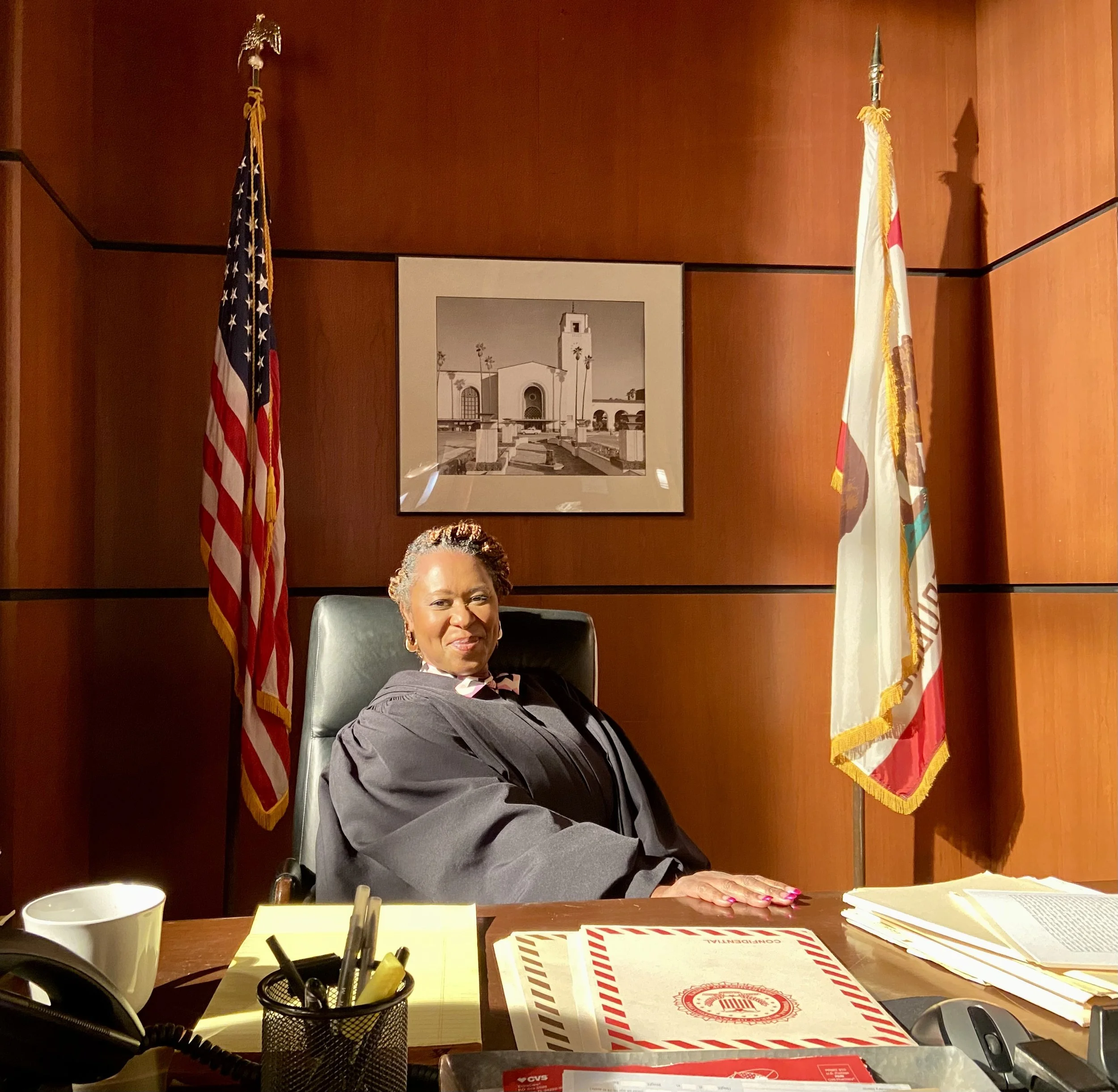 A judge sitting at a desk in a courtroom with flags behind and a photo on the wall.