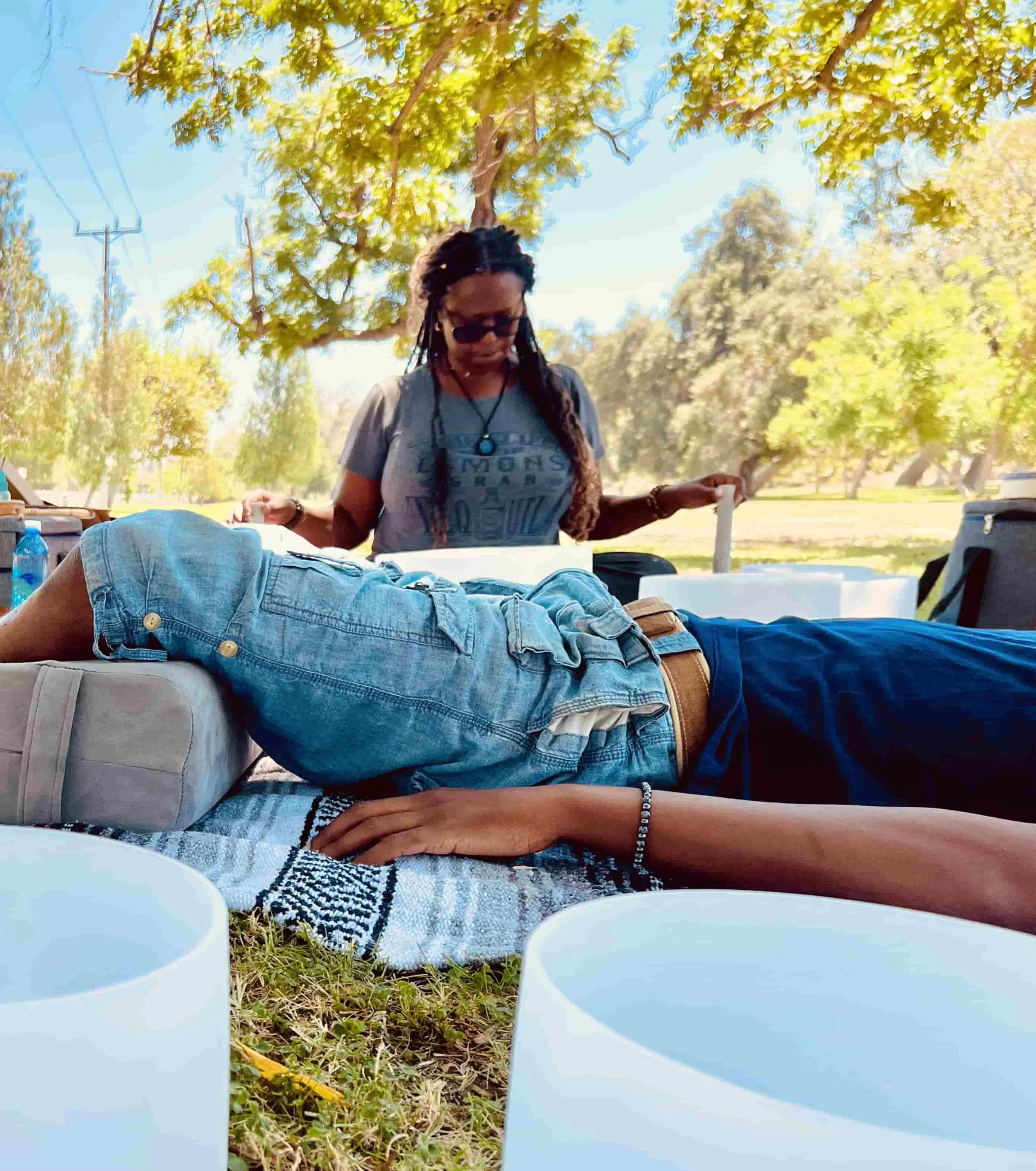 Person lying on a blanket outdoors with another person standing nearby holding sticks, surrounded by trees and instruments.