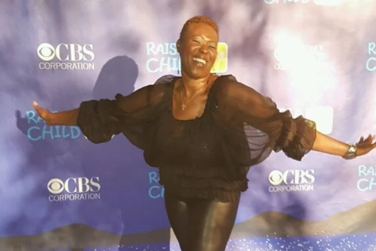 A smiling woman with short hair wearing black clothing, standing with arms outstretched in front of a blue backdrop with CBS Corporation and Raise the Child logos.