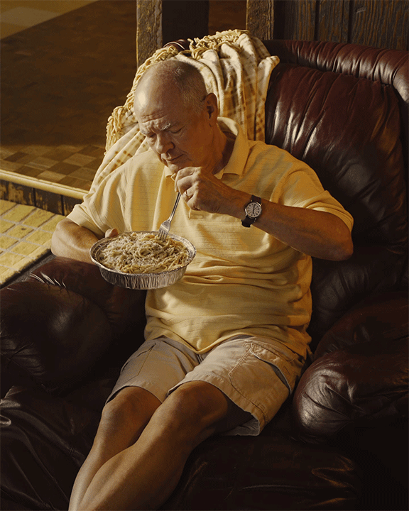 An elderly man sitting on a dark leather armchair, eating from a foil container with a fork.
