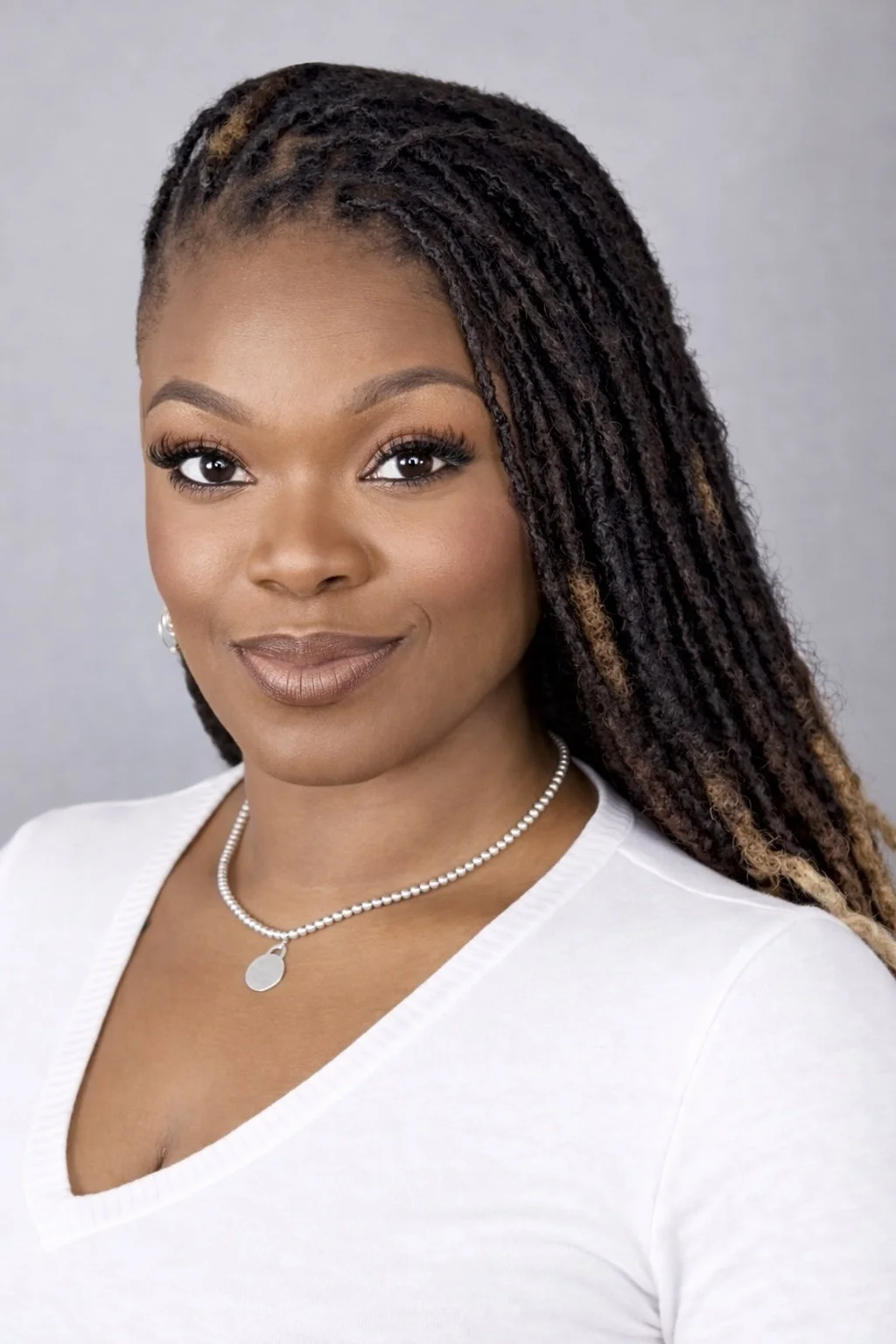 A woman with medium-dark skin, wearing a white top, a pearl necklace, and pearl earrings. She has long, styled dreadlocks with highlights and is smiling, facing the camera against a neutral background.