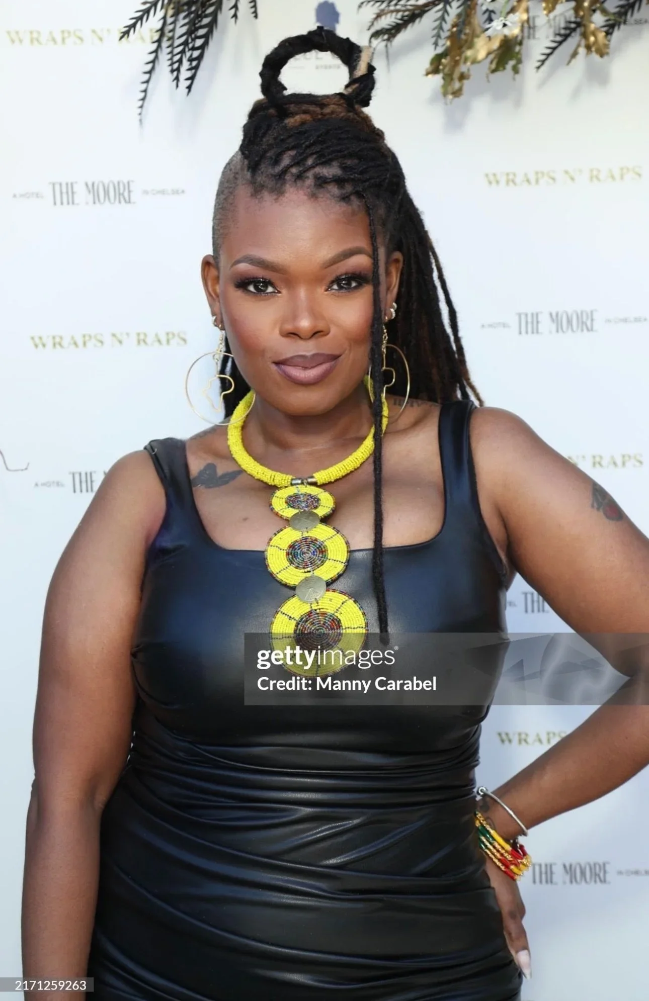 A woman with dark skin and dreadlocks styled in an updo, wearing jewelry and a black sleeveless leather dress, standing in front of a backdrop with the text "WRAPS N' RAPS" and "THE MOORE."