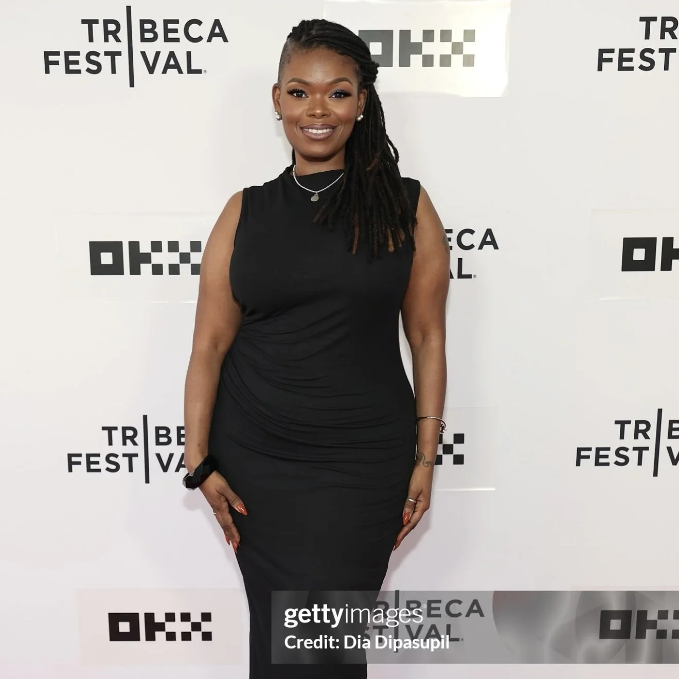 A woman smiling and standing with her hands by her sides in front of a white Tribeca Festival backdrop. She is dressed in a sleeveless black dress with jewelry, including earrings, a necklace, and a bracelet.