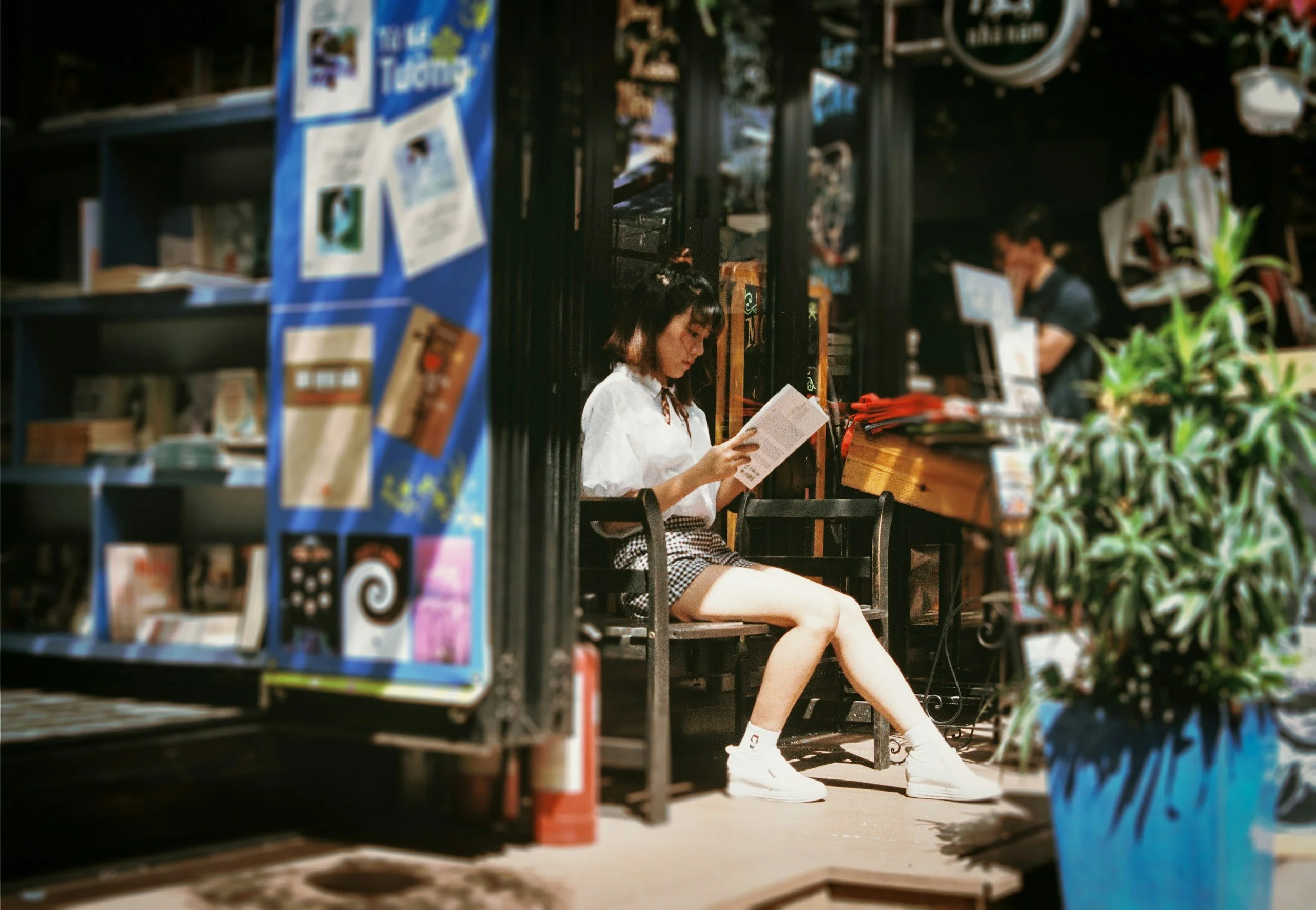 A girl is sitting on a bench reading a book in an outdoor setting. She is wearing a white shirt, plaid shorts, and white sneakers. The scene is seen through a display rack of colorful magazines or books in the foreground.