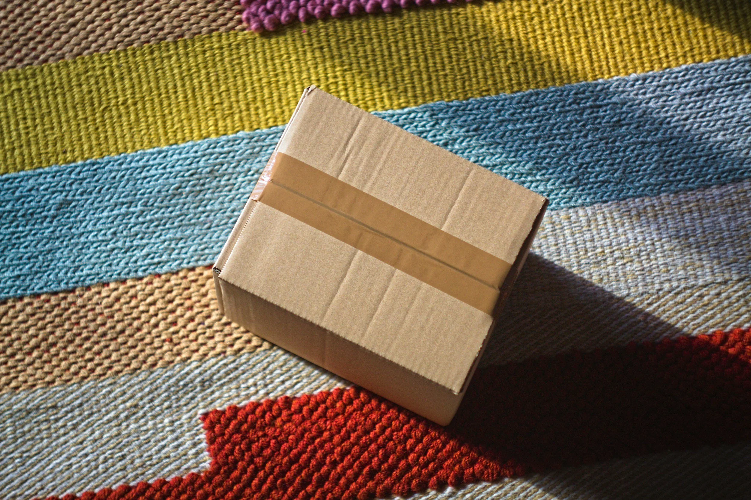 A small cardboard box with tape on it, placed on a colorful woven rug with horizontal strips of various colors including purple, yellow, blue, and red.