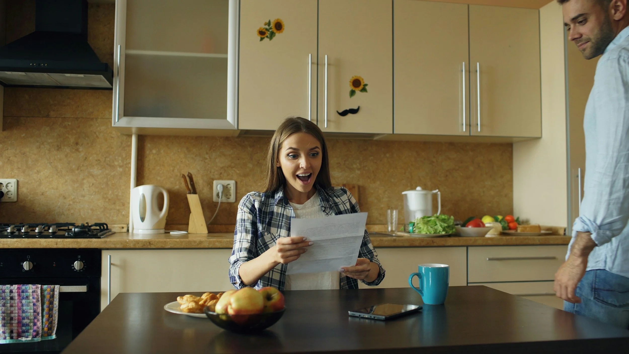 A woman sitting at a kitchen table looking surprised while reading a paper, with a man standing nearby. The table has a bowl of apples, a blue mug, and some pastries. The kitchen has beige cabinets with sunflower decorations and sunflower decals on the cabinet doors, a stovetop, and various kitchen utensils.