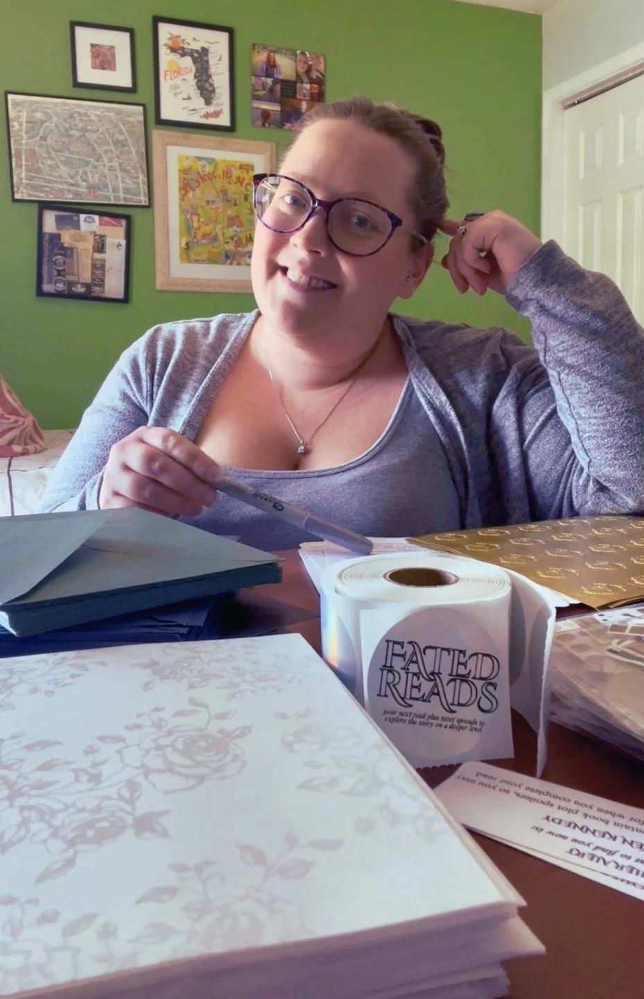 A woman with glasses and a gray shirt sitting at a table with papers, a pen, and a booklet labeled 'EATED READS' in a room with green walls decorated with framed pictures and posters.