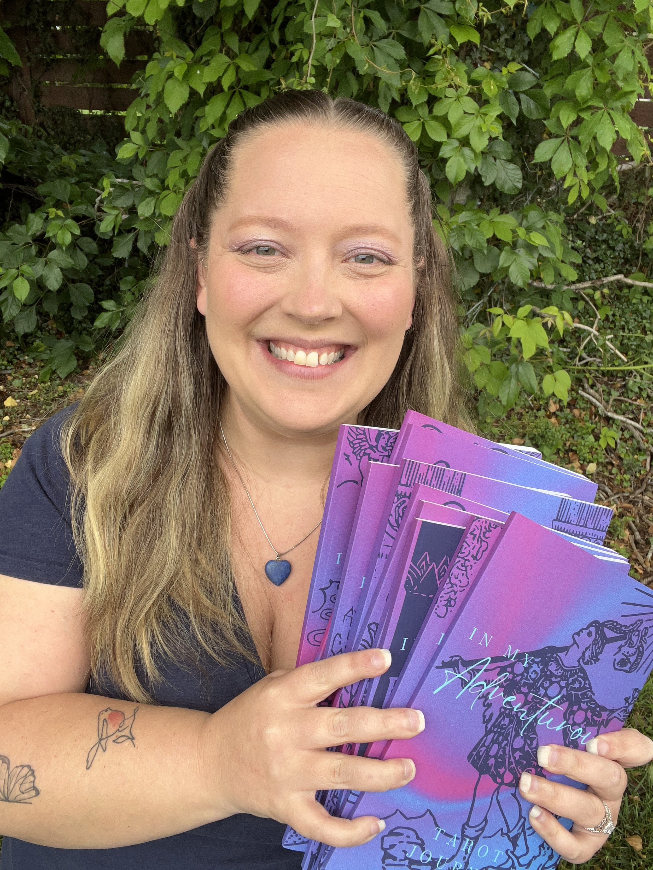 A woman with long, wavy blonde hair smiling and holding colorful tarot journals in front of green leafy bushes. She has a tattoo of a butterfly on her forearm and is wearing a blue heart-shaped necklace and a silver ring.