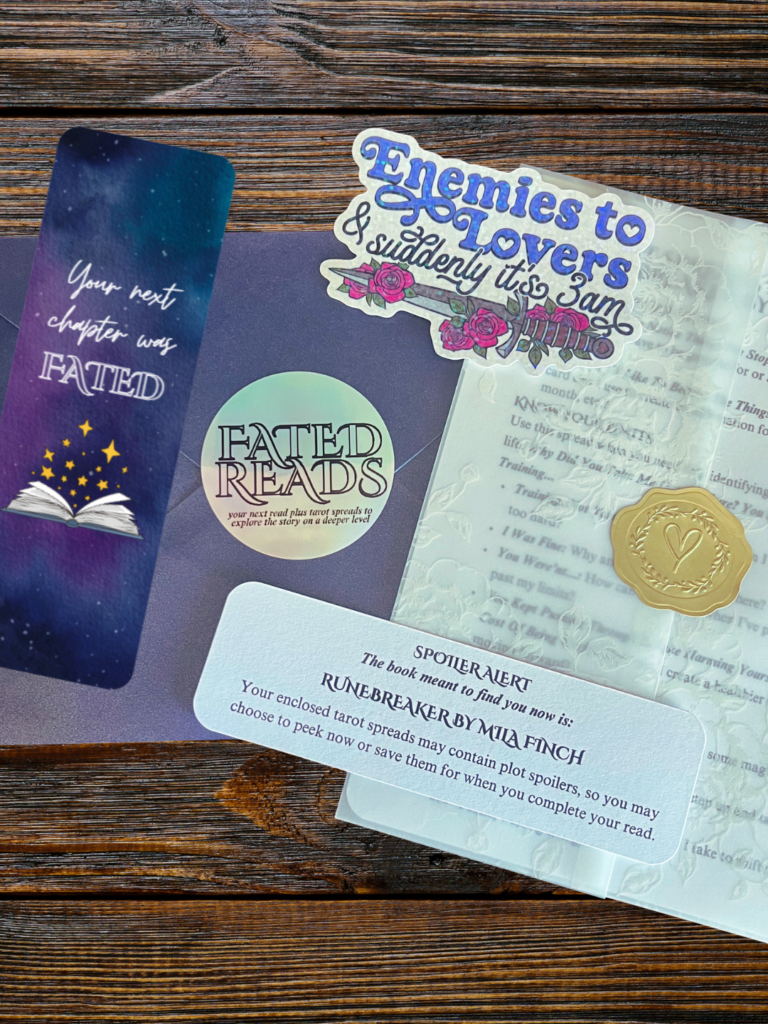 A wooden table with tarot reading materials, including a sticker with the text "Enemies to Lovers & suddenly it's 3 am," a lapel pin with a heart, a booklet titled "Fated Reads," and a spoiler alert note about a book called "Runebreaker" by Mia Finch.
