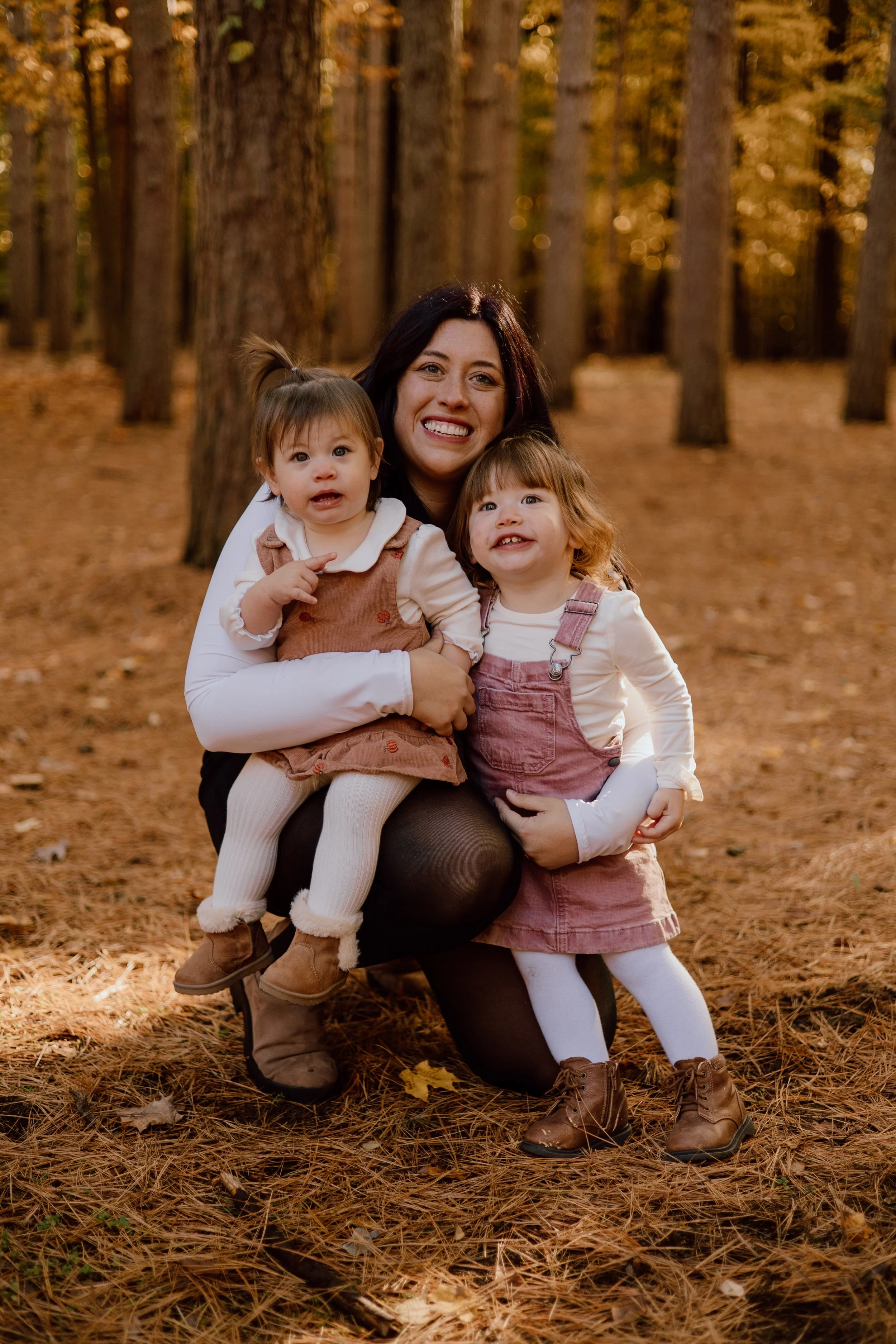 A woman with two young girls in a forest during fall, surrounded by trees and fallen leaves.