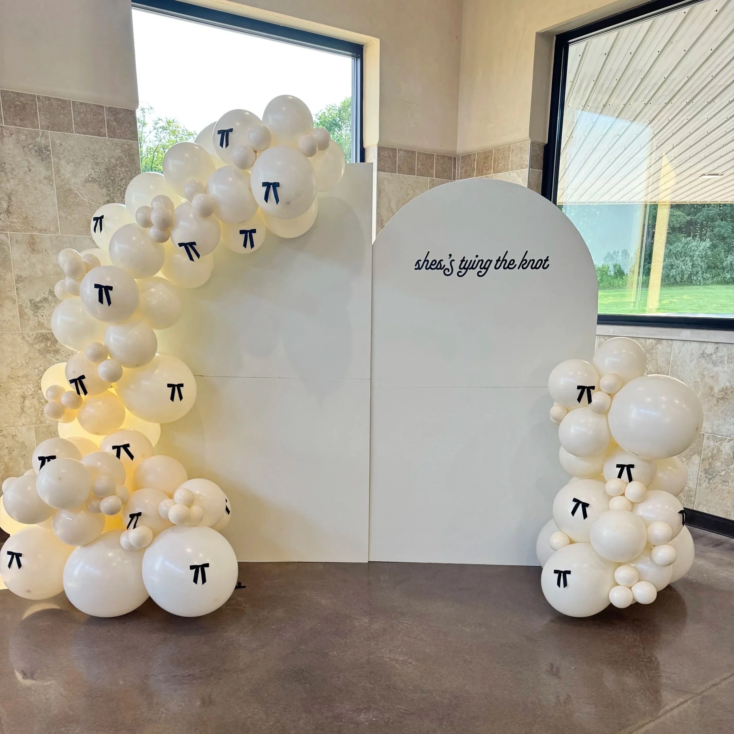 A balloon display with white balloons arranged around a white backdrop, decorated with black pi symbol. The backdrop has a sign that reads "she's tying the knot." The balloons are of various sizes with some featuring the pi symbol.