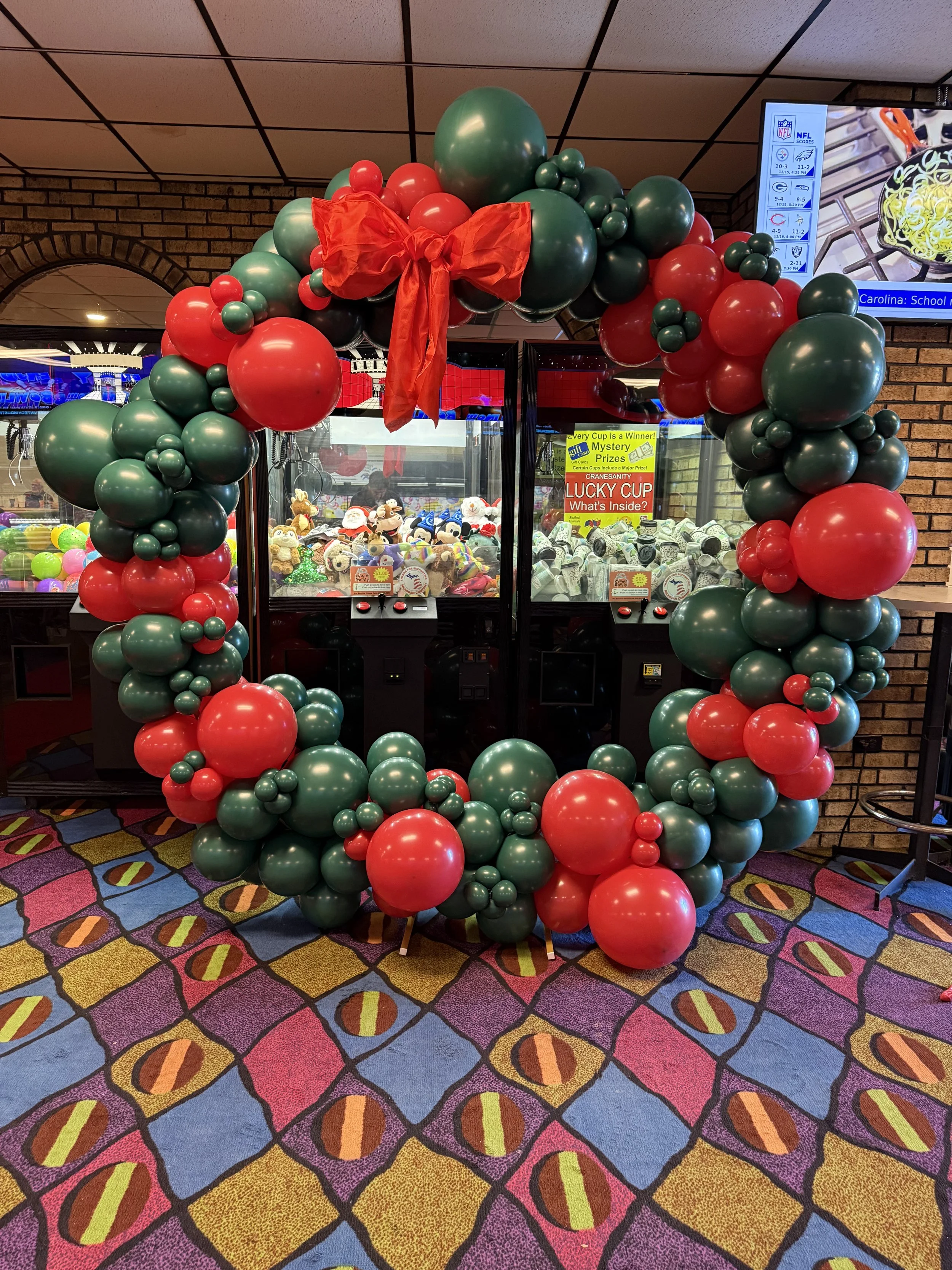 A large Christmas-themed balloon wreath with red and green balloons, decorated with a red bow, surrounds a claw machine filled with plush toys at an arcade.