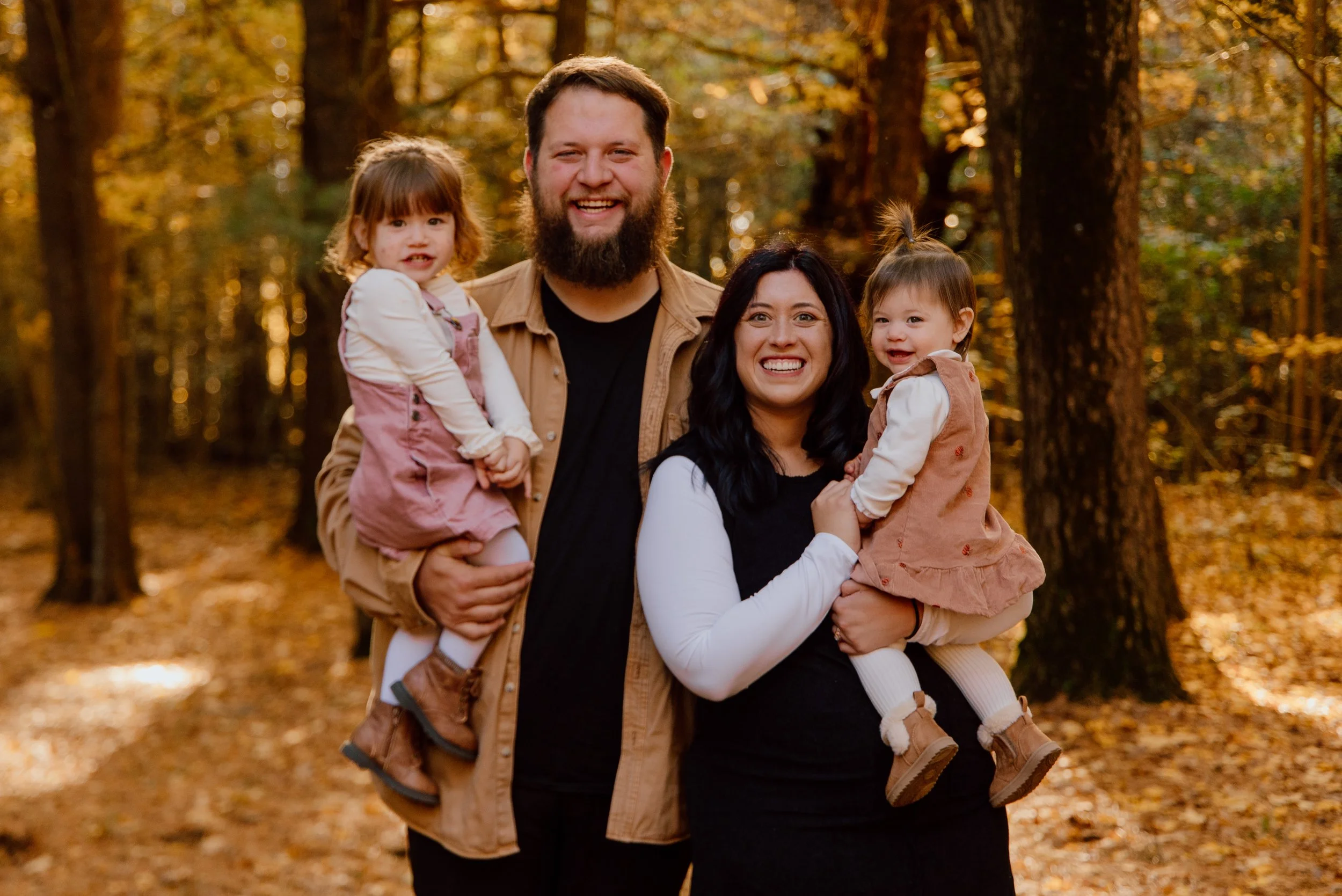 A family of four enjoying time outdoors in a park with autumn foliage. The father has a beard, wearing a beige jacket and black shirt. The mother has black hair, wearing a black dress with white sleeves, and is holding a young girl with a brown dress. The father is holding another young girl with red hair wearing a pink dress. All members are smiling.
