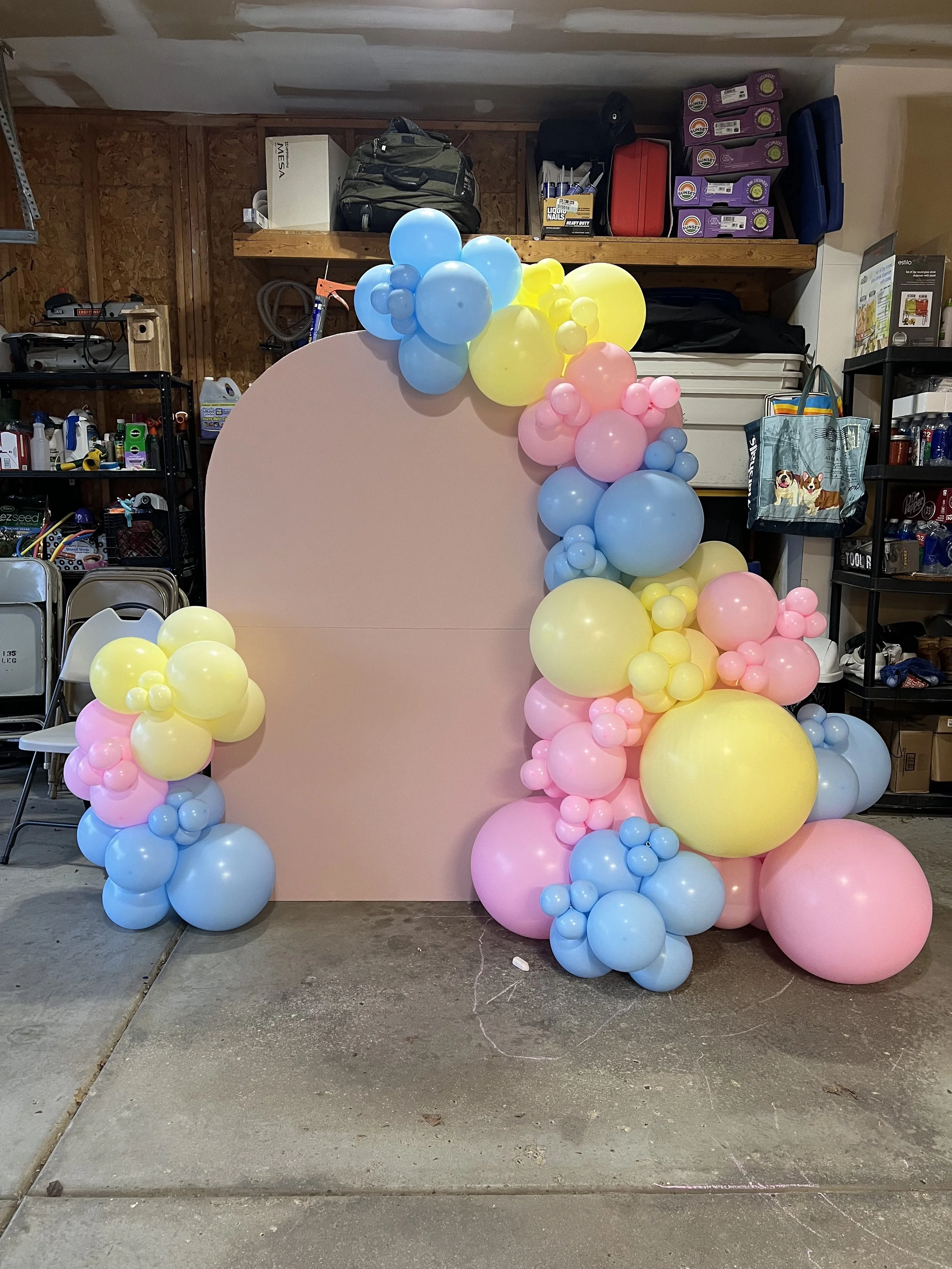A pastel pink backdrop decorated with colorful balloon clusters in blue, yellow, pink, and purple, set up in a garage or storage room.