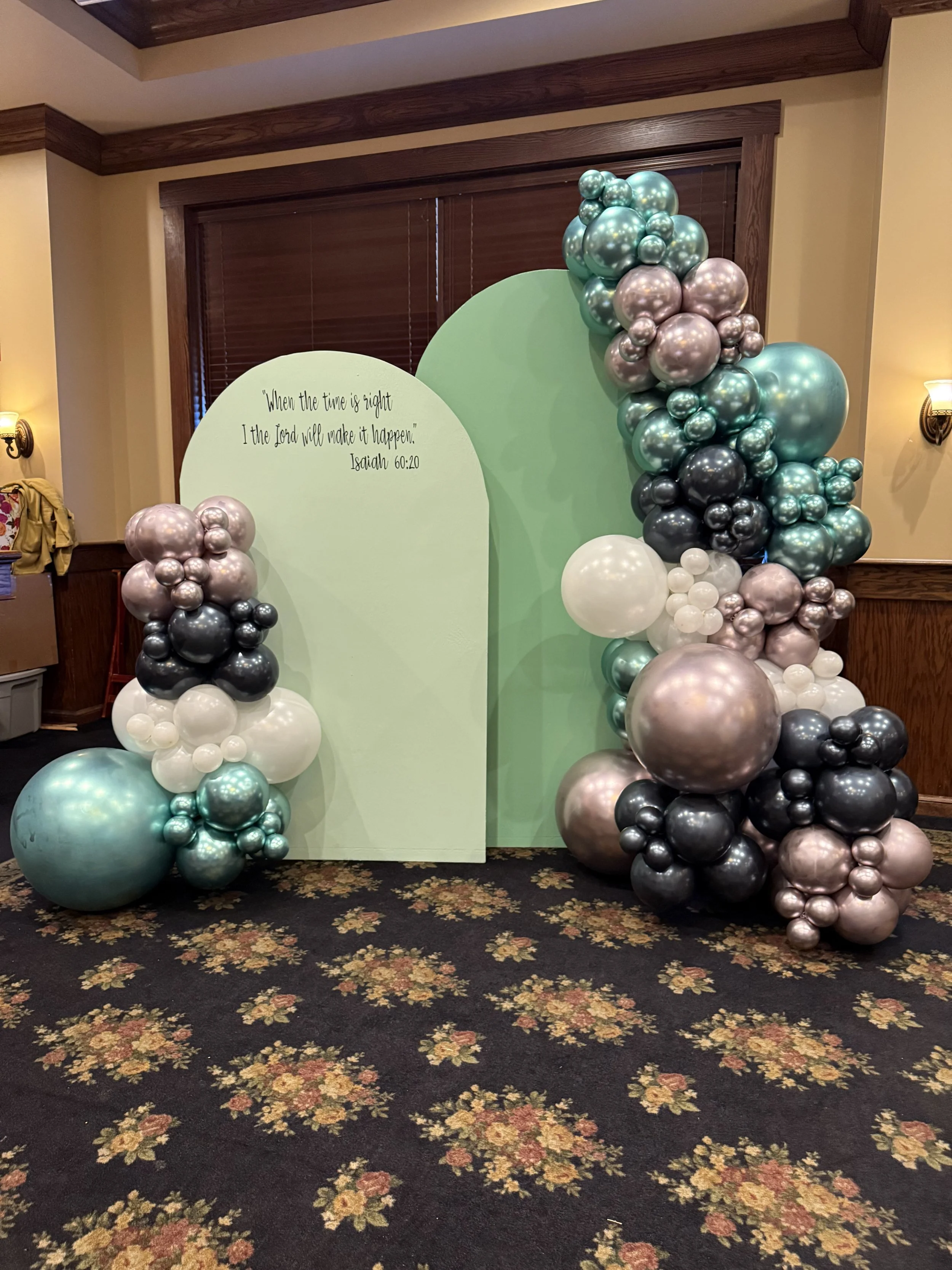 Decorative backdrop with balloons in shades of teal, black, silver, and white, with a green cutout shape featuring a Bible verse from Isaiah 60:20, in a room with floral carpet and wooden window frames.