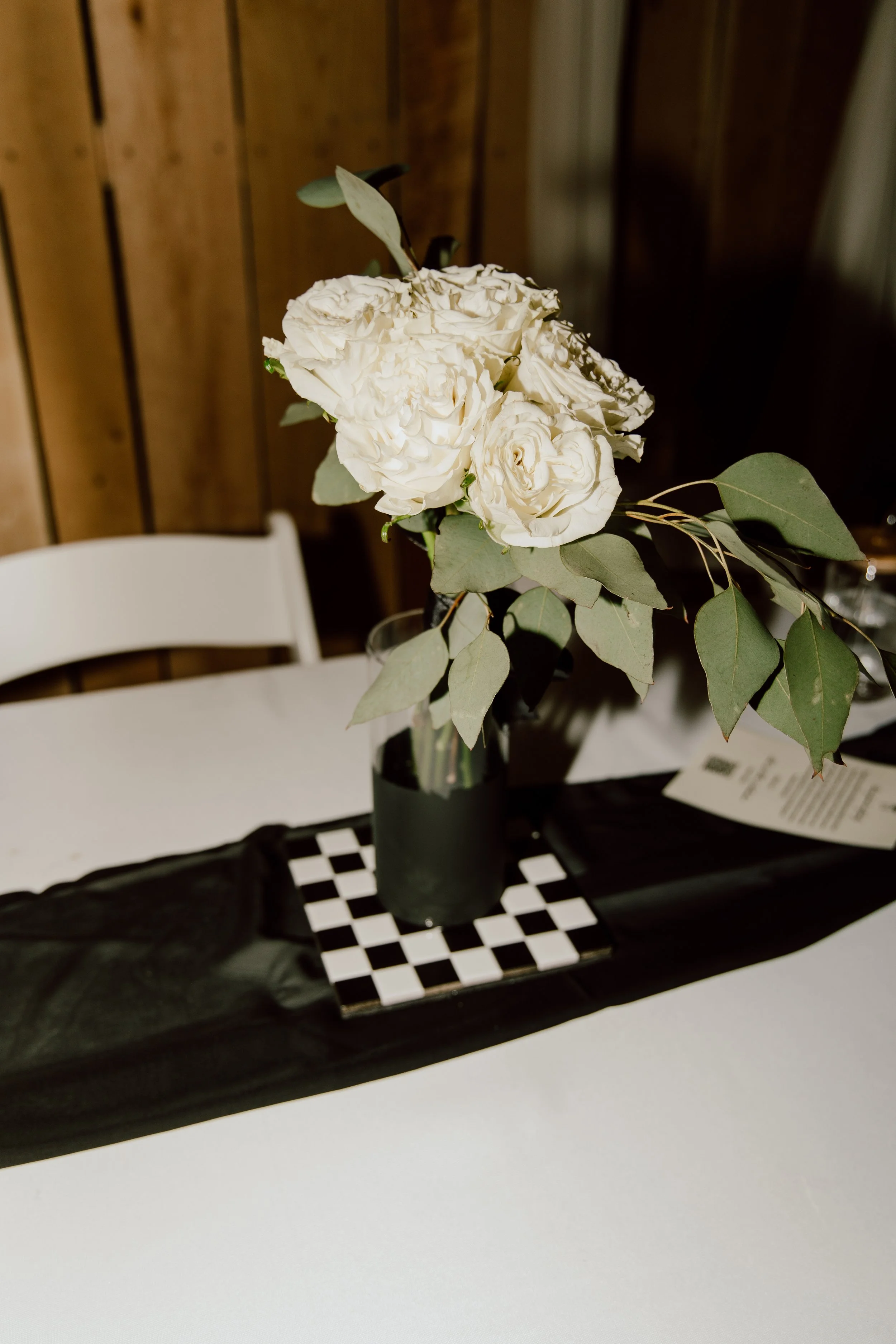A bouquet of white flowers and green leaves in a black vase on a checkered table mat, placed on a white table with a black table runner.