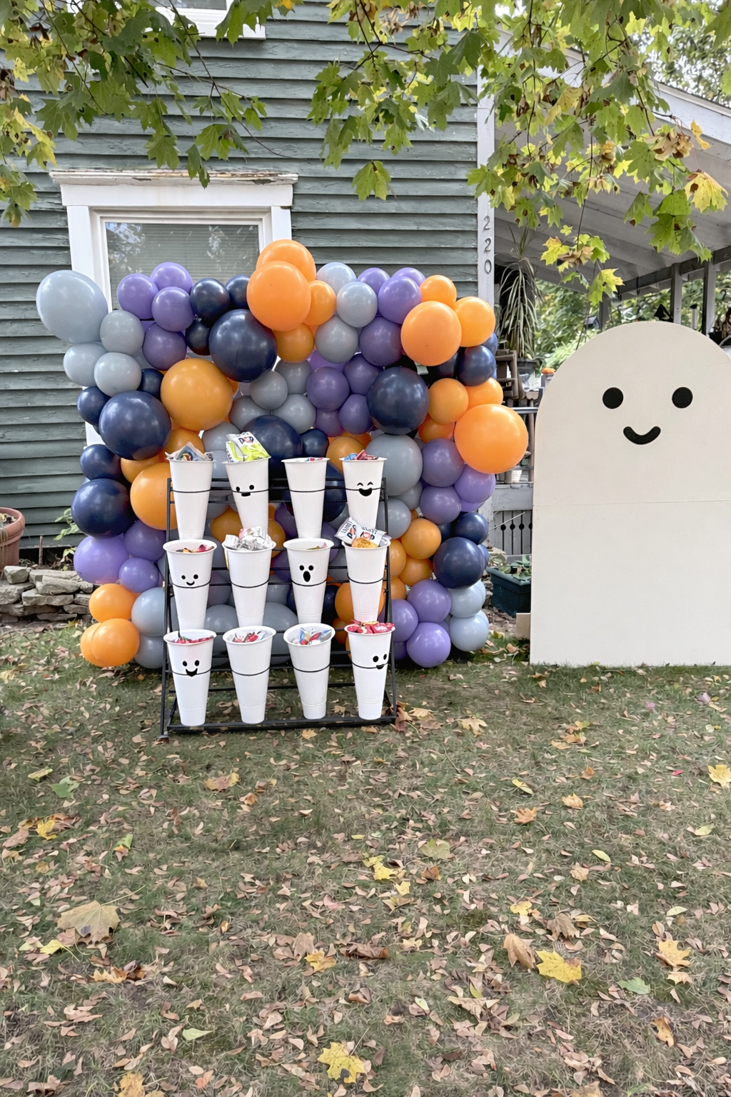 A Halloween-themed outdoor display with a balloon arch in purple, orange, black, and gray around a white ghost figure with a smiling face. In front, there is a black metal stand holding several white cups with cute black smiley faces, filled with can