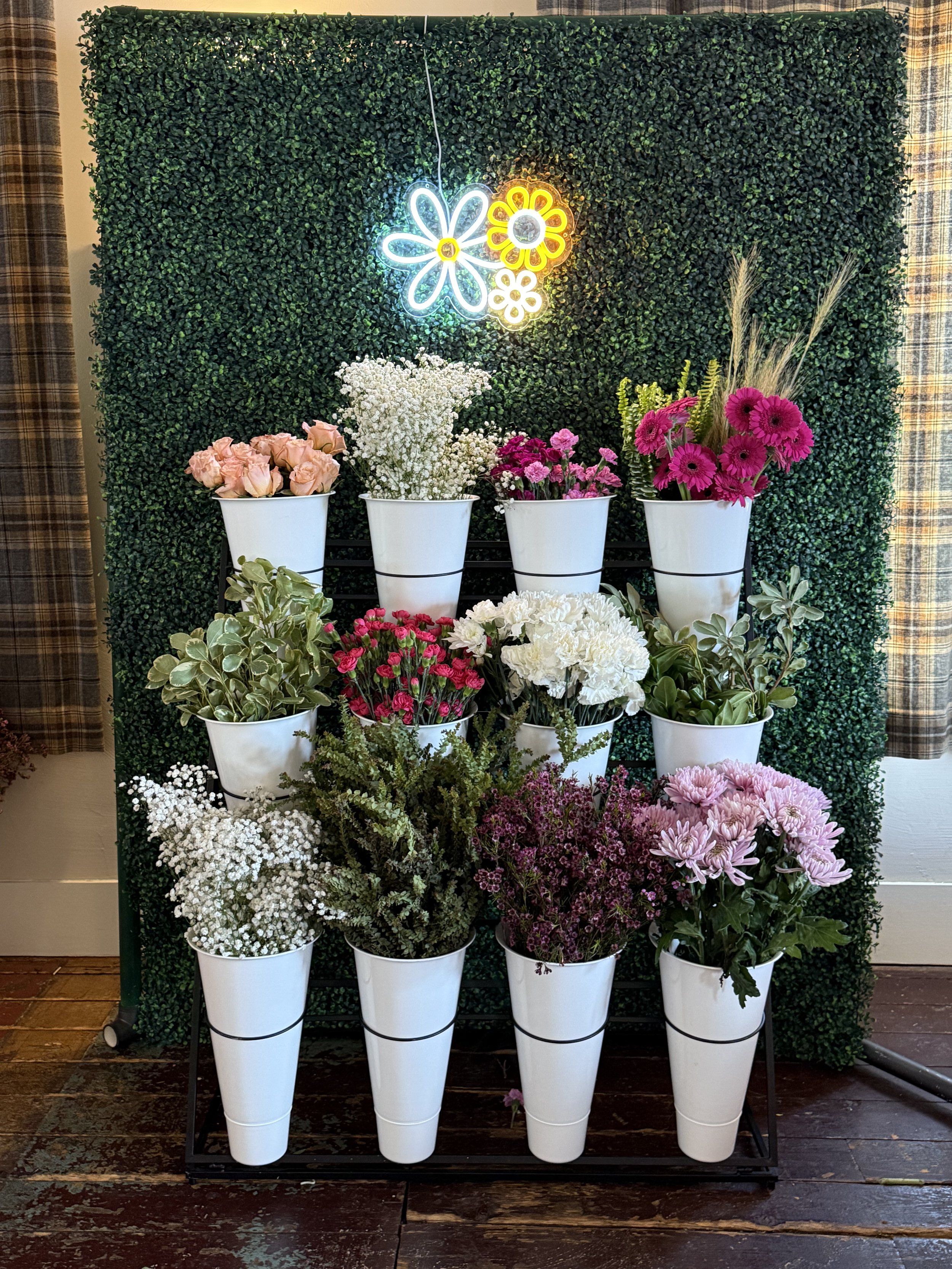 Display of various fresh flowers in white pots on a black metal stand against a green leafy wall, with neon flower signs above.