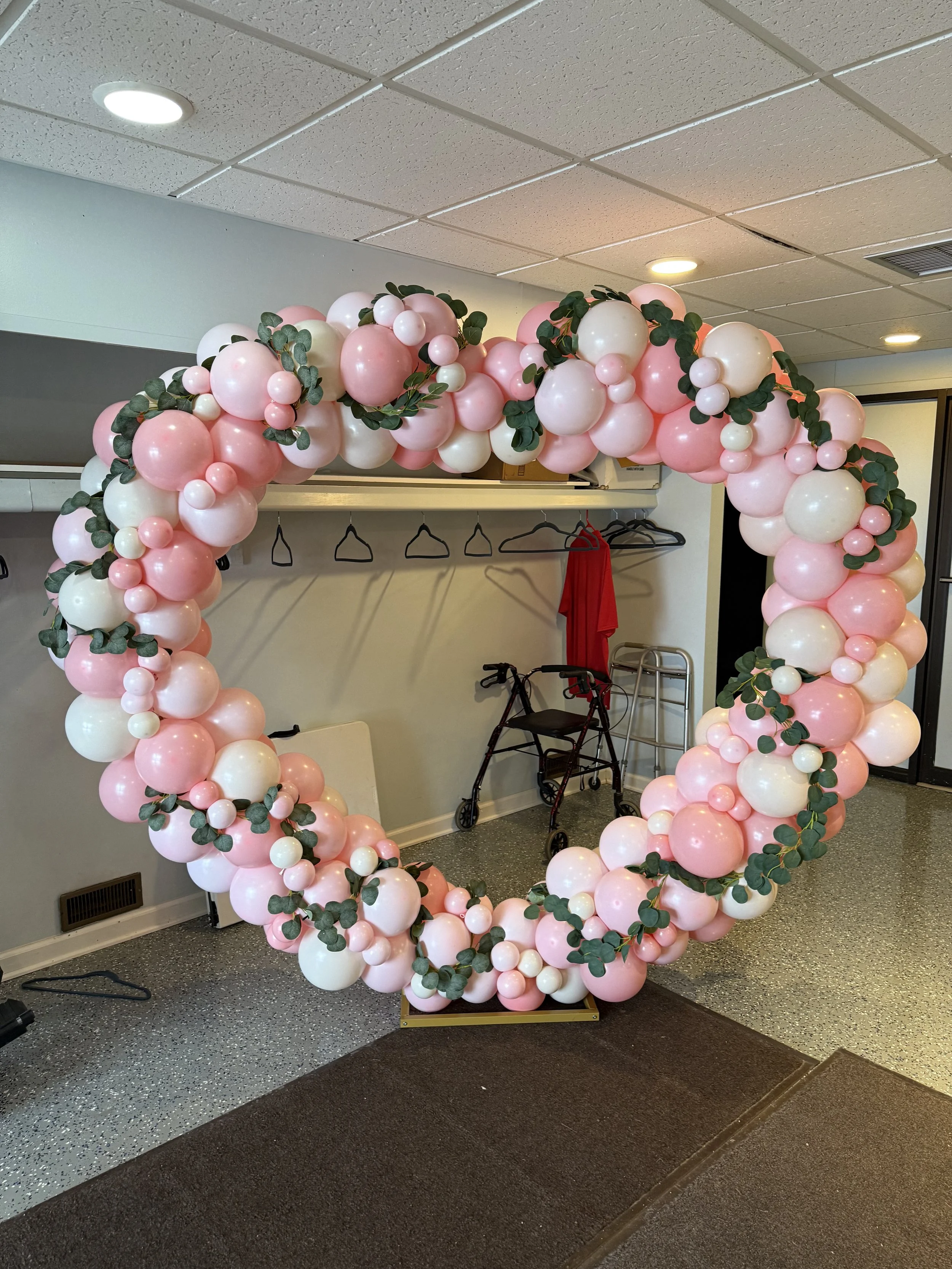 A decorative floral balloon arch with pink and white balloons and green leaves, set up indoors on a carpeted floor, near a wall with a coat rack and mobility aids.