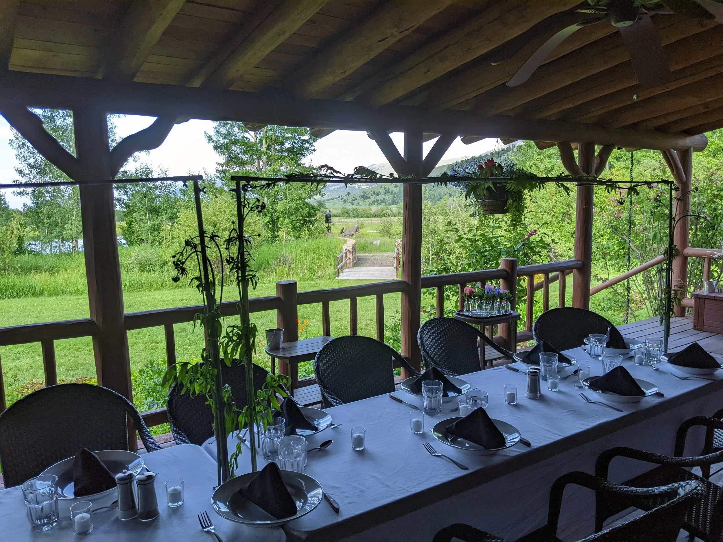 A covered outdoor patio area with a view of lush green trees and fields in the distance. The patio has a wooden roof and railing, with a table set for a meal with glasses, black napkins, and plates.