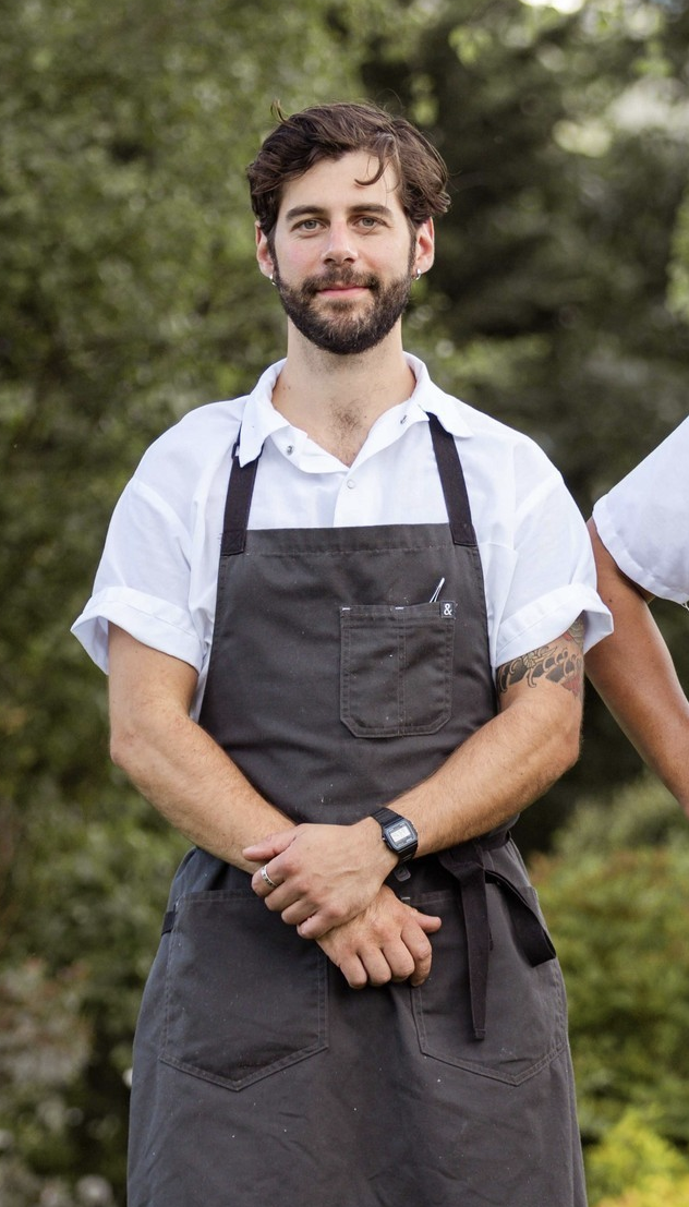 A man with brown hair and a beard, wearing a white shirt and black apron, standing outdoors with greenery in the background.