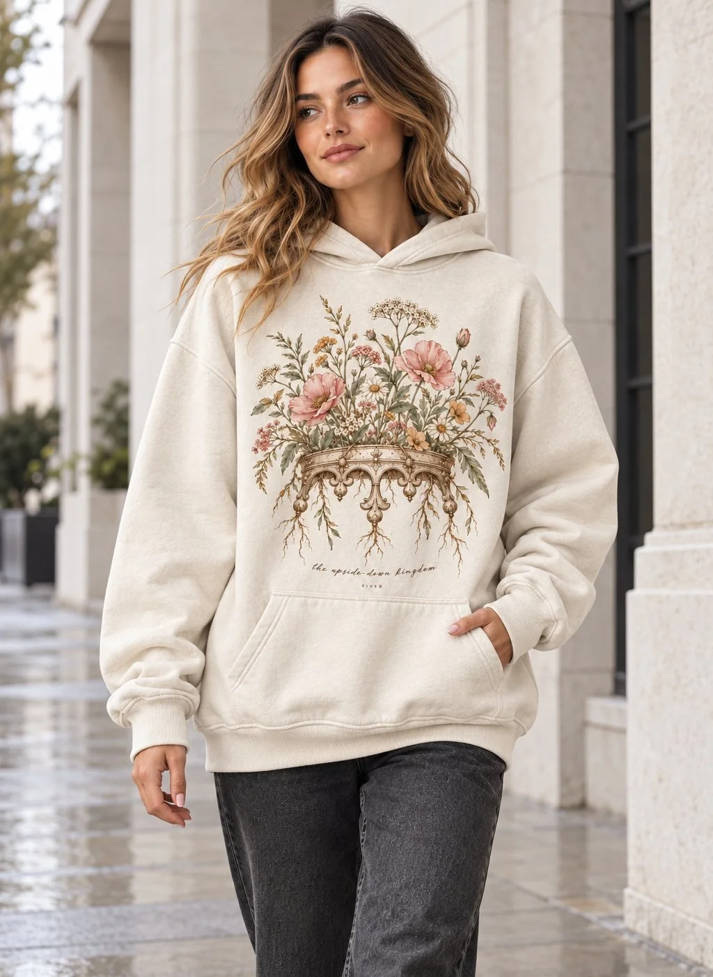 A woman in a cream hoodie printed with a floral crown turned upside down, flowers above and roots hanging below, with the script line the upside-down kingdom and a small FIVE8 mark