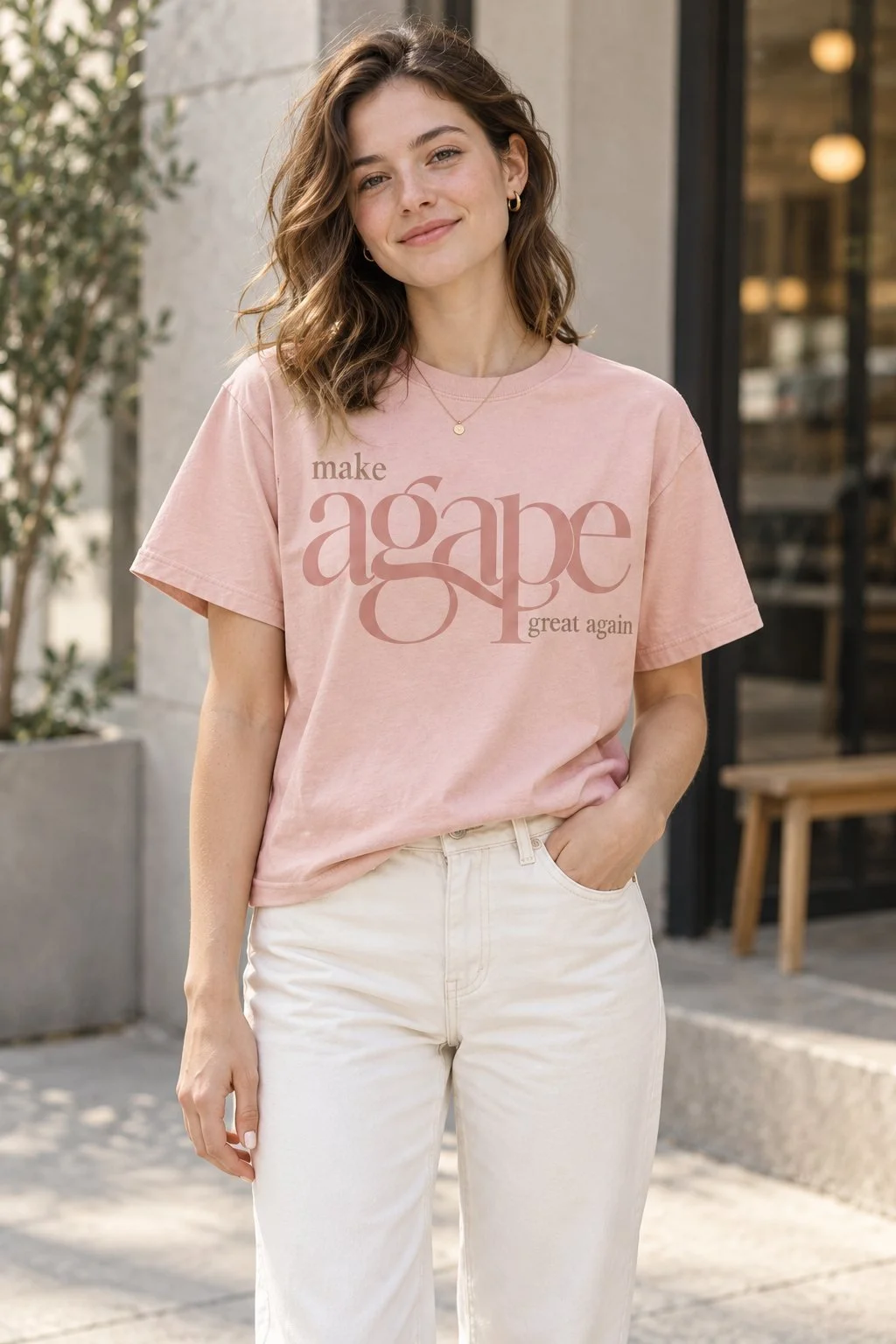 A woman in a soft pink tee printed with make agape great again, the word agape set large in a tonal serif, standing in front of a stone wall and a cafe window