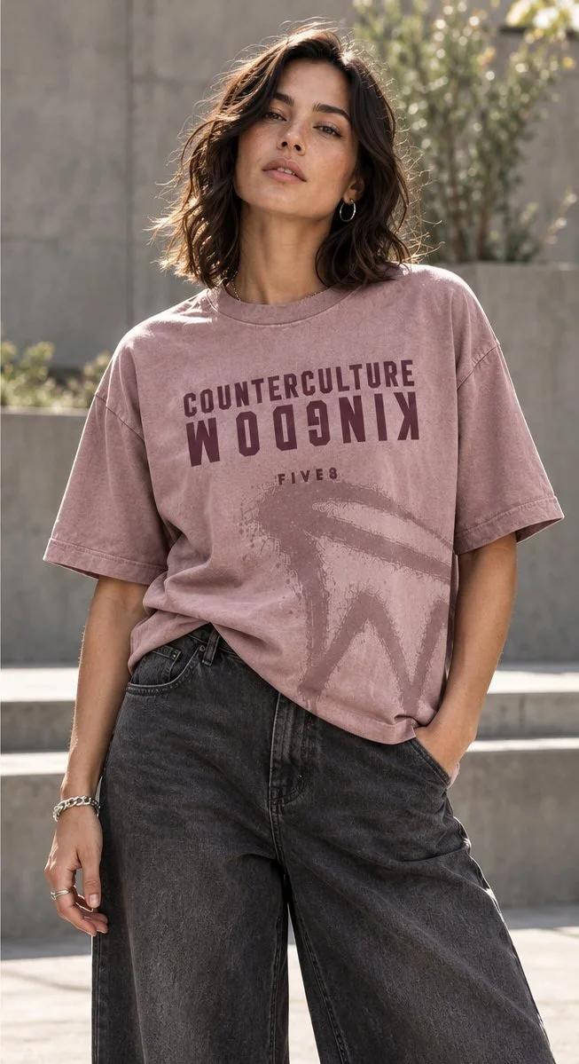 A woman in a mauve oversized tee printed in plum with COUNTERCULTURE above an upside-down KINGDOM and a sprayed mark below