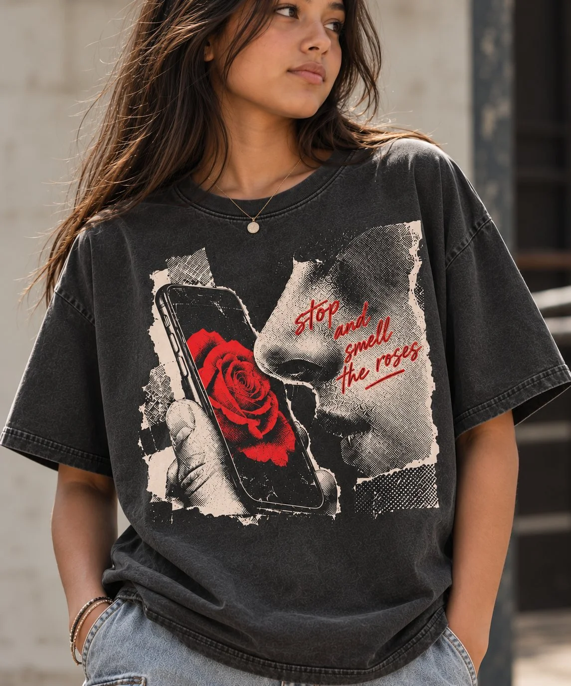 A woman in a washed black tee printed with a torn-paper halftone collage of a hand holding a phone with a red rose, a nose beside it and the red script stop and smell the roses