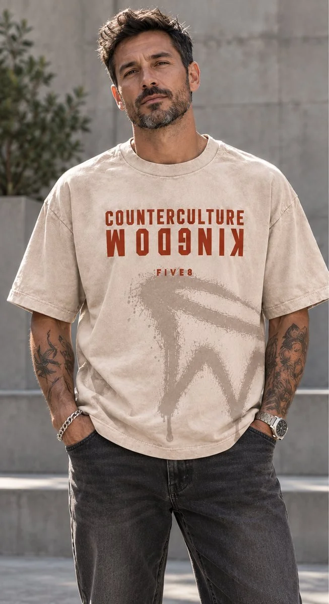 A man in a beige oversized tee printed in rust with COUNTERCULTURE above an upside-down KINGDOM and a sprayed mark below