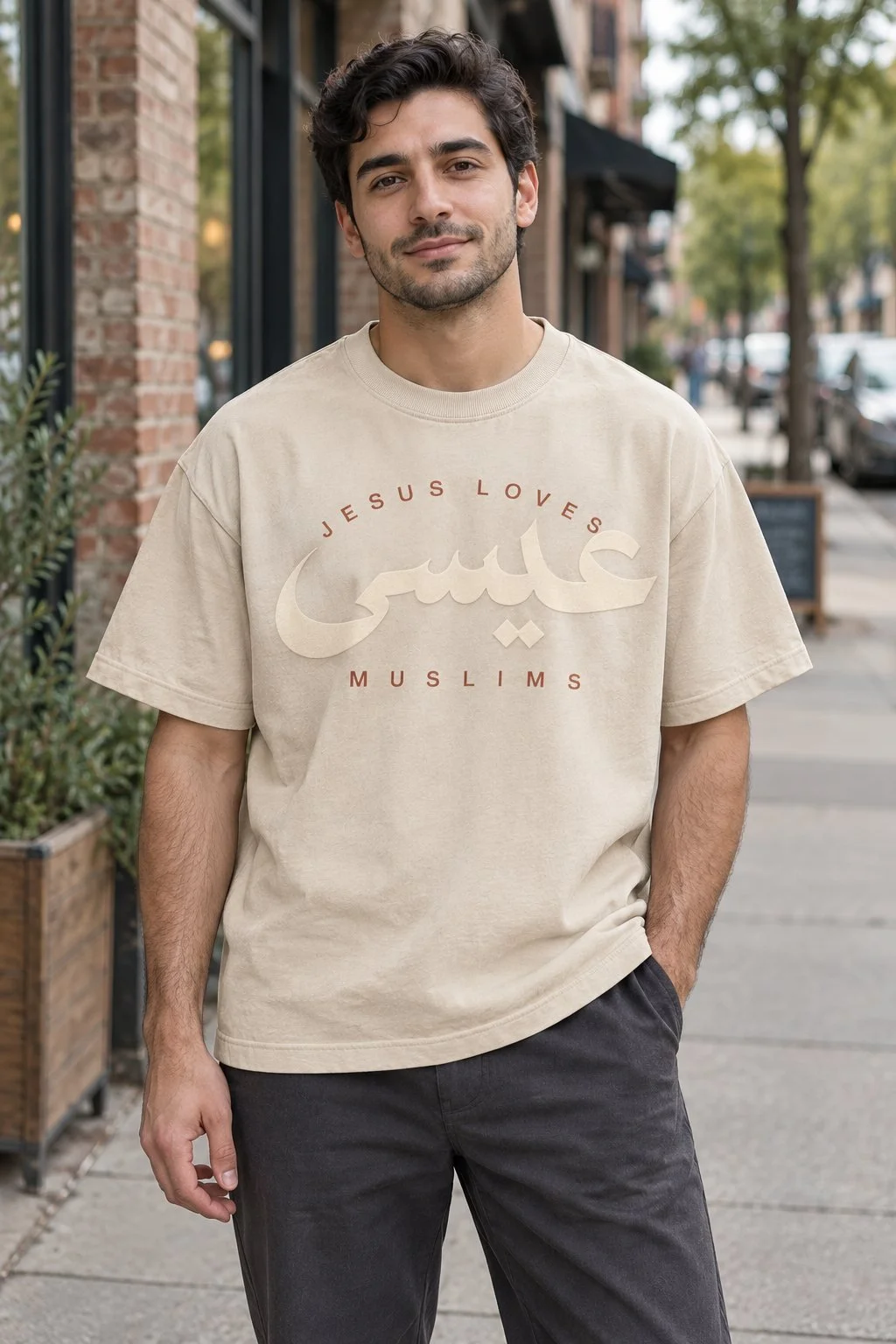 A man in a beige oversized tee printed with JESUS LOVES above the name Jesus in Arabic calligraphy and MUSLIMS below, standing on a brick-lined city sidewalk