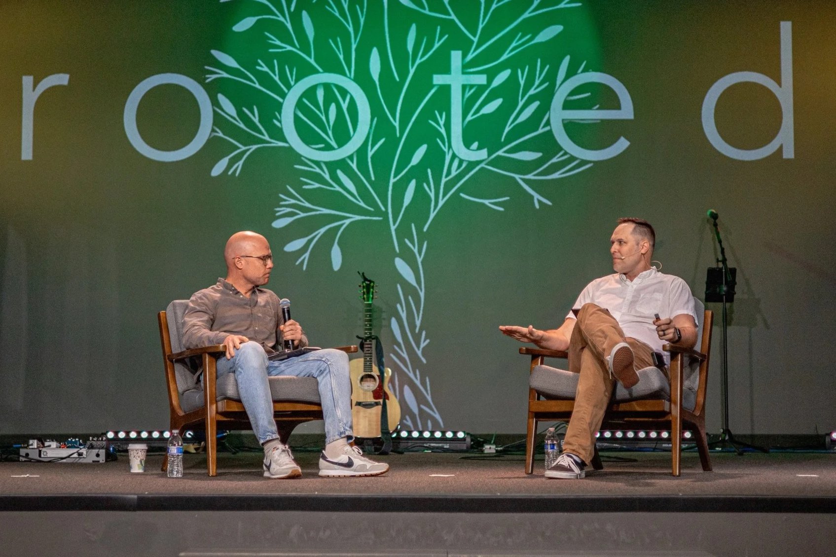 Two men participating in a discussion on a stage. The man on the left, bald and wearing glasses, is holding a microphone and sitting in a chair. The man on the right, with short hair, is gesturing with his hand. Behind them is a large green backdrop with a white tree design and the word 'rooted' written across it. There are microphones, water bottles, a guitar, and stage equipment visible on the stage.